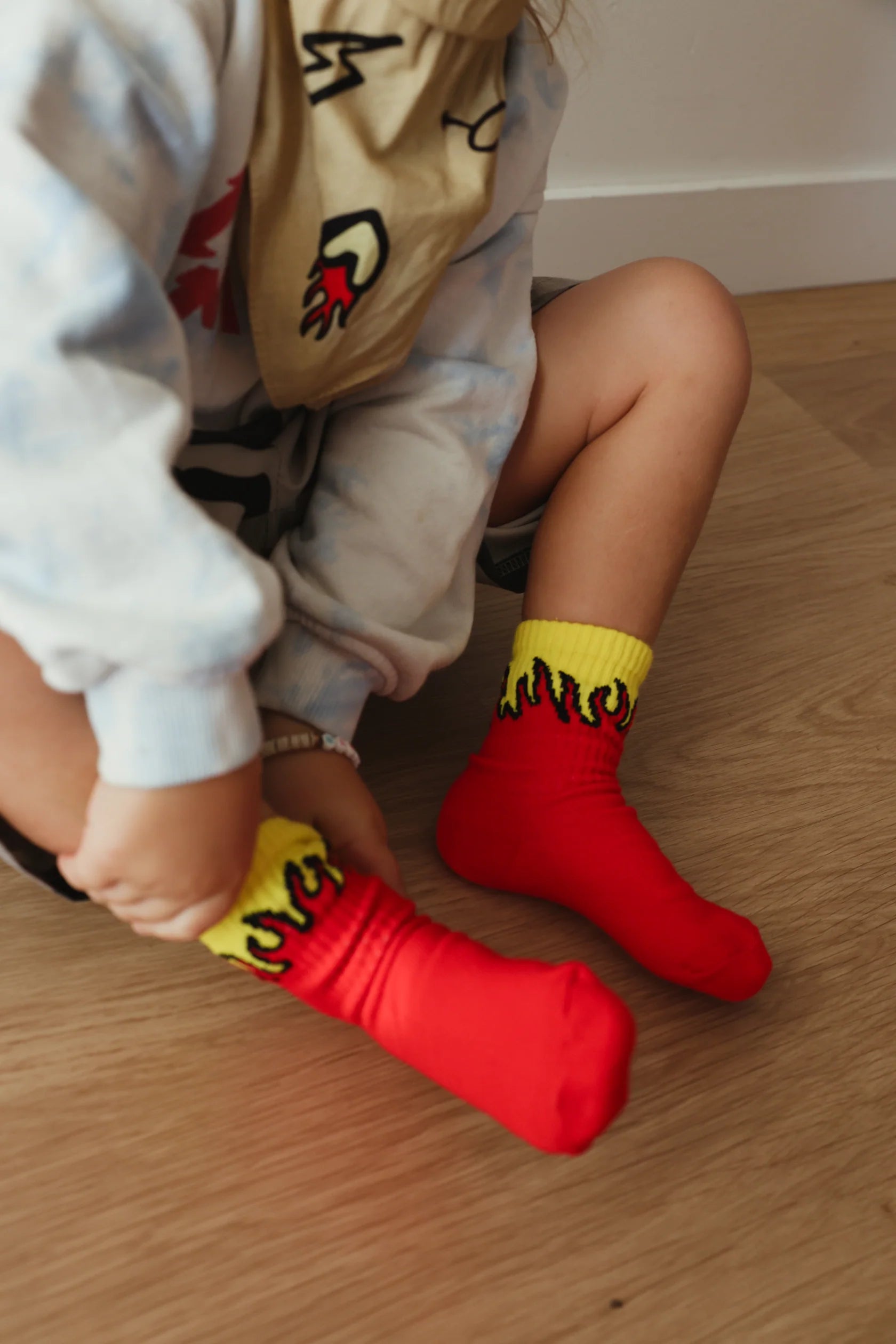 A close-up of a light-skinned child sitting on a wooden floor, wearing a white sweatshirt and putting on TINY LOVE CLUB's Fire Socks. The socks are bright red with yellow and black flame patterns at the top and feature elastic banded ankles for a snug fit. The child is also wearing a bib adorned with red and black graphics.