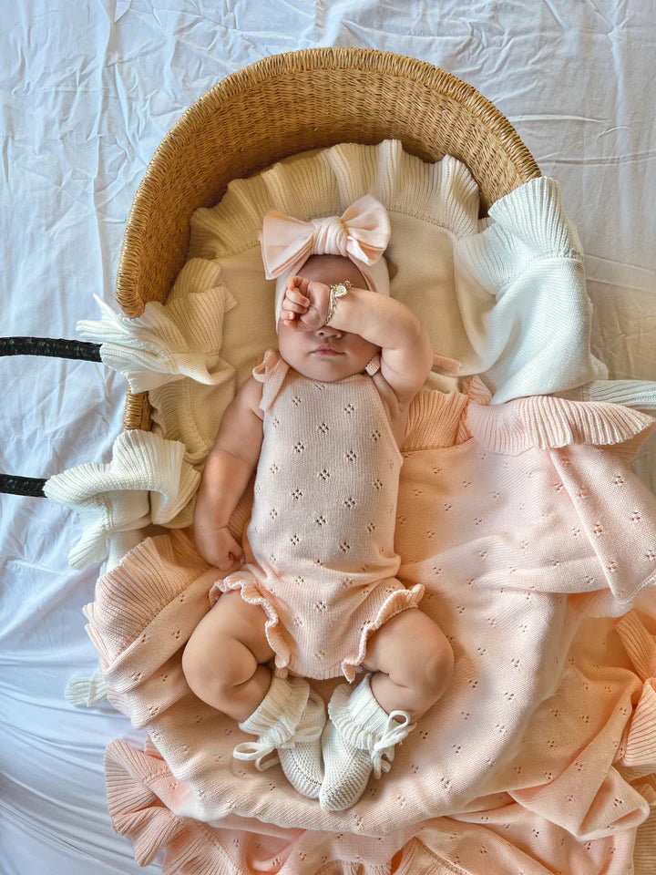 A baby lies in a wicker bassinet with a white ruffled mattress. The baby, dressed in a light pink Frill Bodysuit Primrose from ZIGGY LOU's Signature Collection, along with matching booties and a large bow headband, has one arm resting on their face. The background is a white sheet.