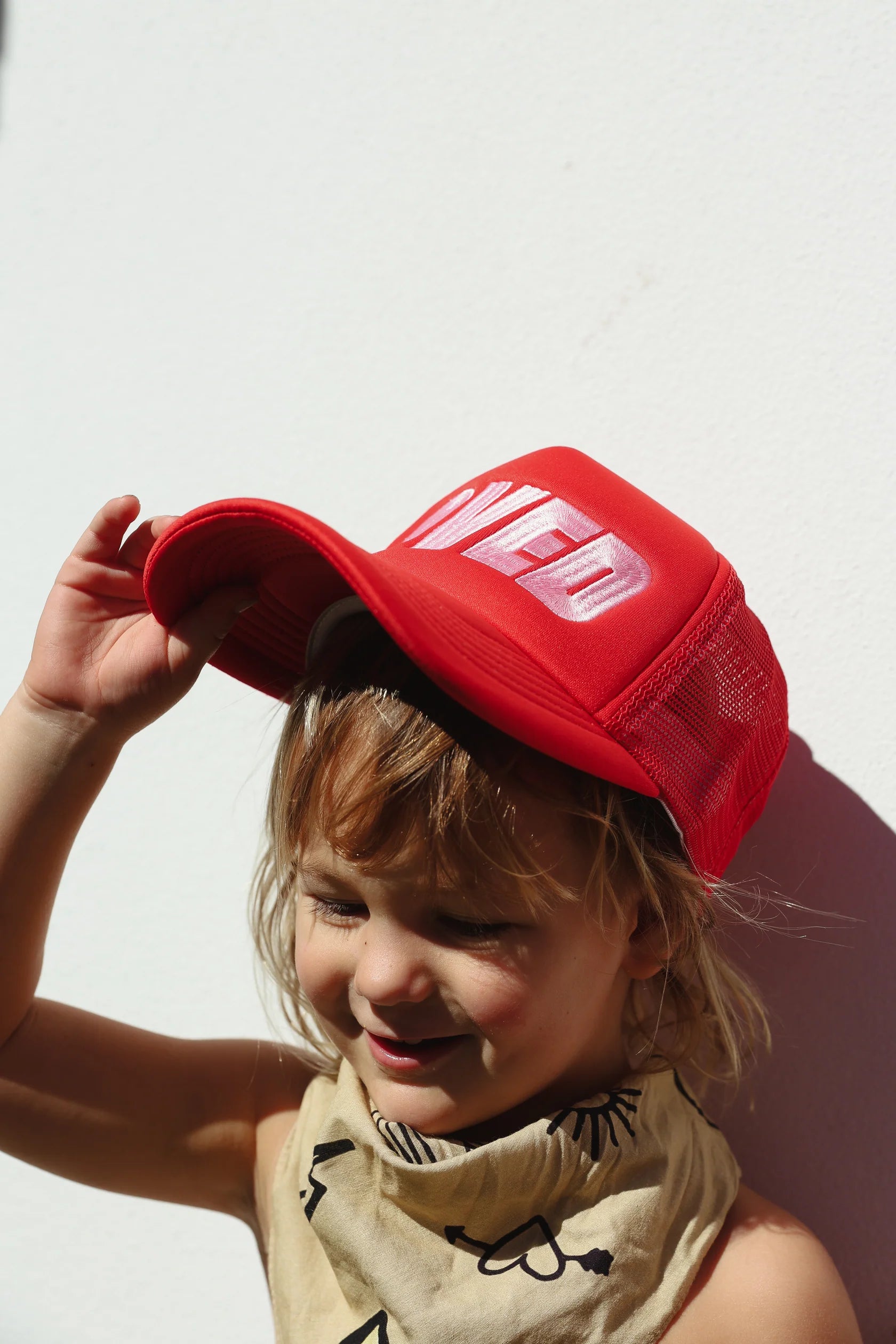 A smiling young child wears the TINY LOVE CLUB's "So Loved Trucker Cap," featuring an adjustable strap for comfort. The red cap with "LOVED" text pairs perfectly with a light-patterned bib against a white background, while sunlight casts a clear shadow on the wall.