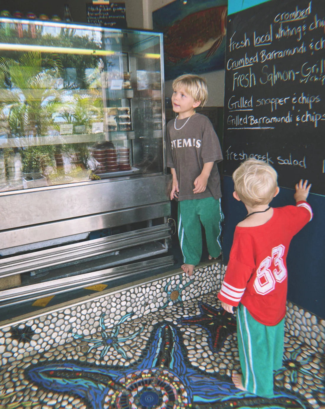 Two children eagerly gaze at a food display in a shop. The older one wears a brown "ARTEMIS" shirt, while the younger dons ZIGGY ZAZA's Twin Stripe Terry Pant Verde with an elastic waist and a red "83" shirt. The floor displays a colorful mosaic design featuring a starfish.