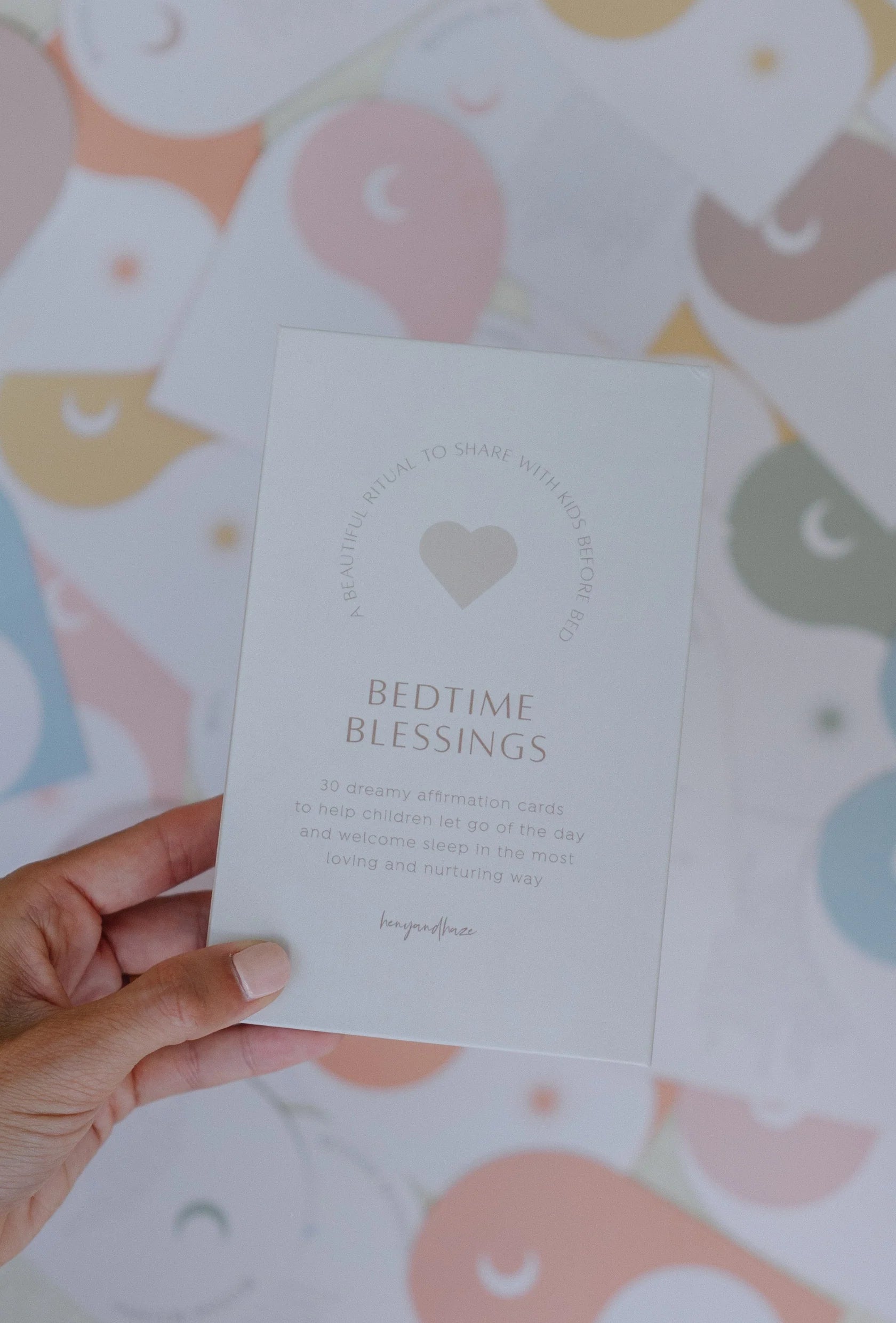 A hand holding a card titled "Bedtime Blessings" from HENY AND HAZE, featuring a heart illustration. The card mentions 30 affirmation cards designed to help children tackle sleep challenges. In the background, various cards from the Bedtime Blessings Deck are displayed, showcasing pastel-colored designs with stars and moons.