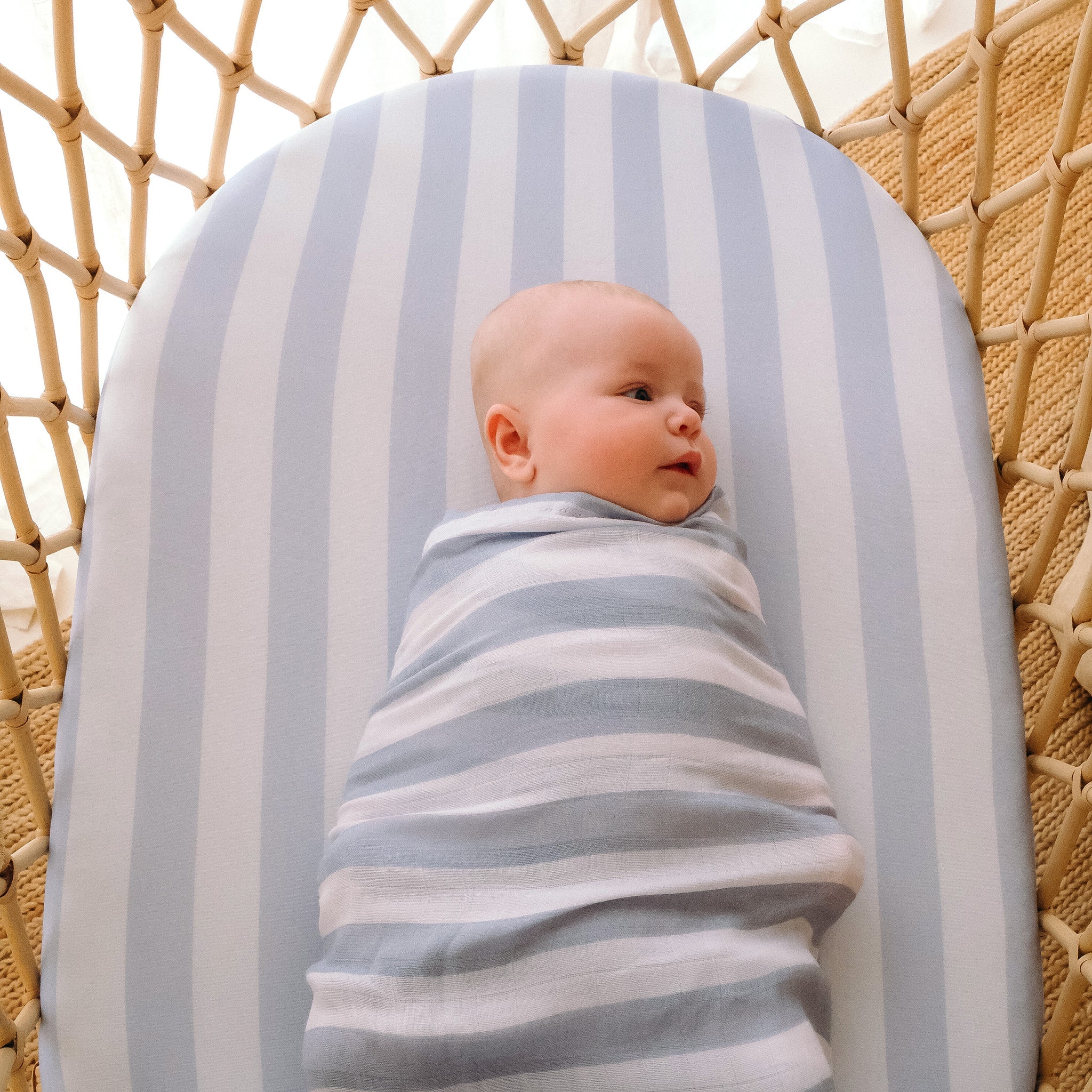 A baby lies on the FOX & FALLOW - Cloud Stripe Organic Fitted Bassinet Sheet/Change Pad Cover in a woven bassinet, wrapped in a matching blue and white striped blanket.