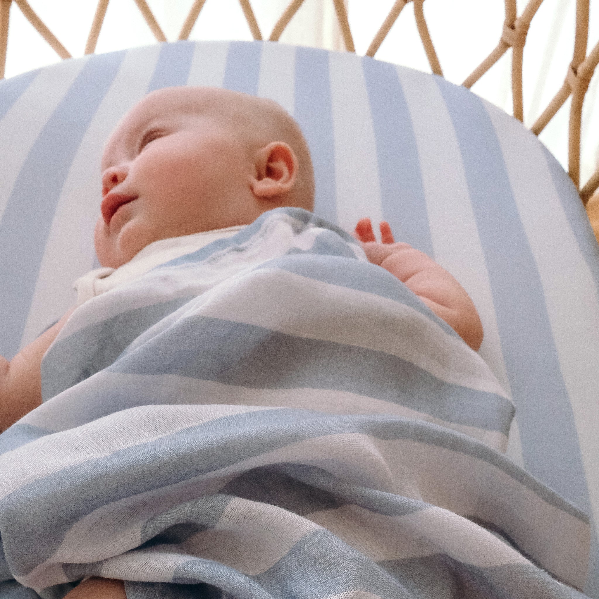 A baby rests on the FOX & FALLOW Cloud Stripe Organic Fitted Bassinet Sheet/Change Pad Cover, featuring light blue and white stripes for gentle comfort and style.