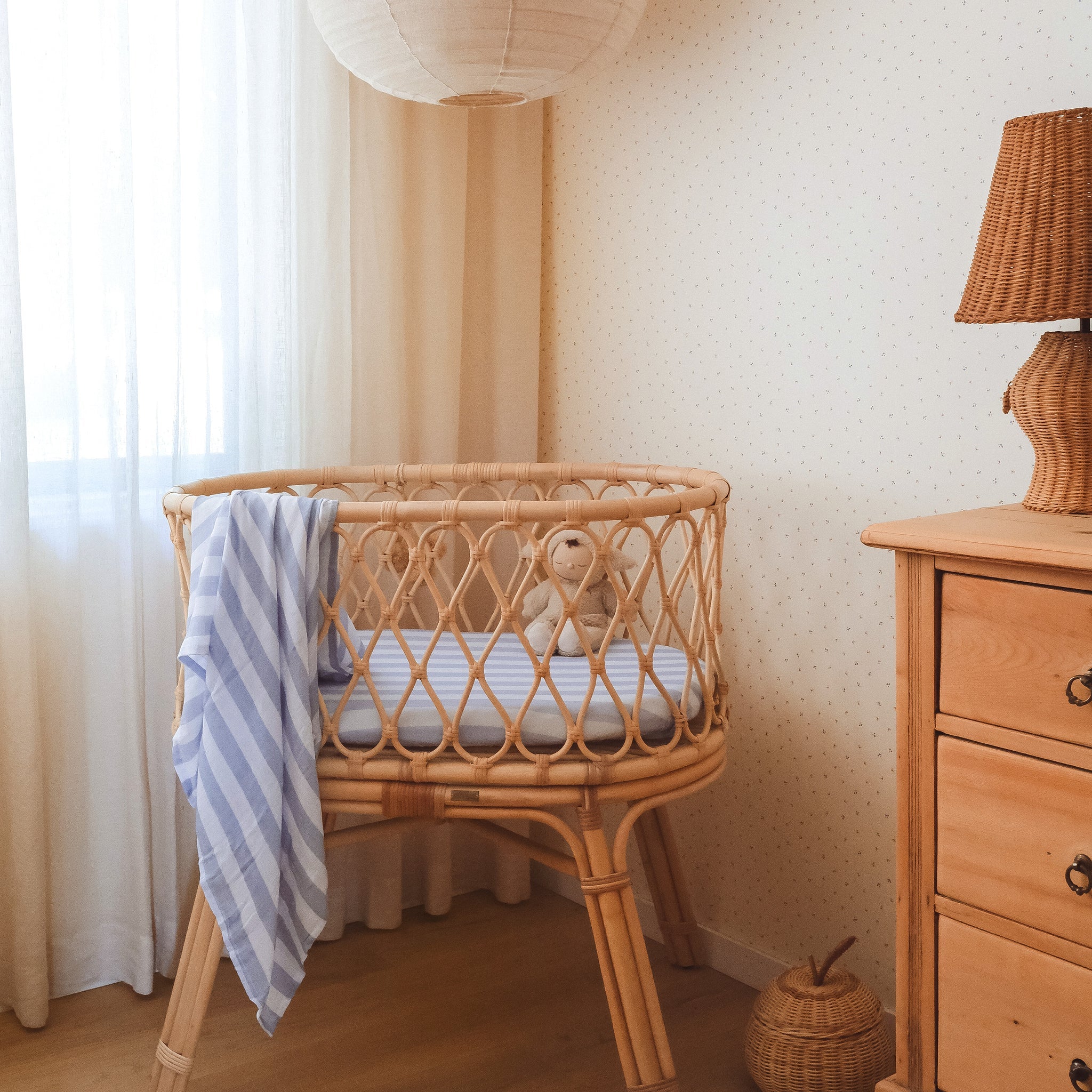 A cozy nursery with a round rattan crib featuring the FOX & FALLOW Cloud Stripe Organic Fitted Bassinet Sheet, a blue and white striped blanket, plush toy, woven lamp, and wooden dresser—all in warm, natural tones with soft lighting.