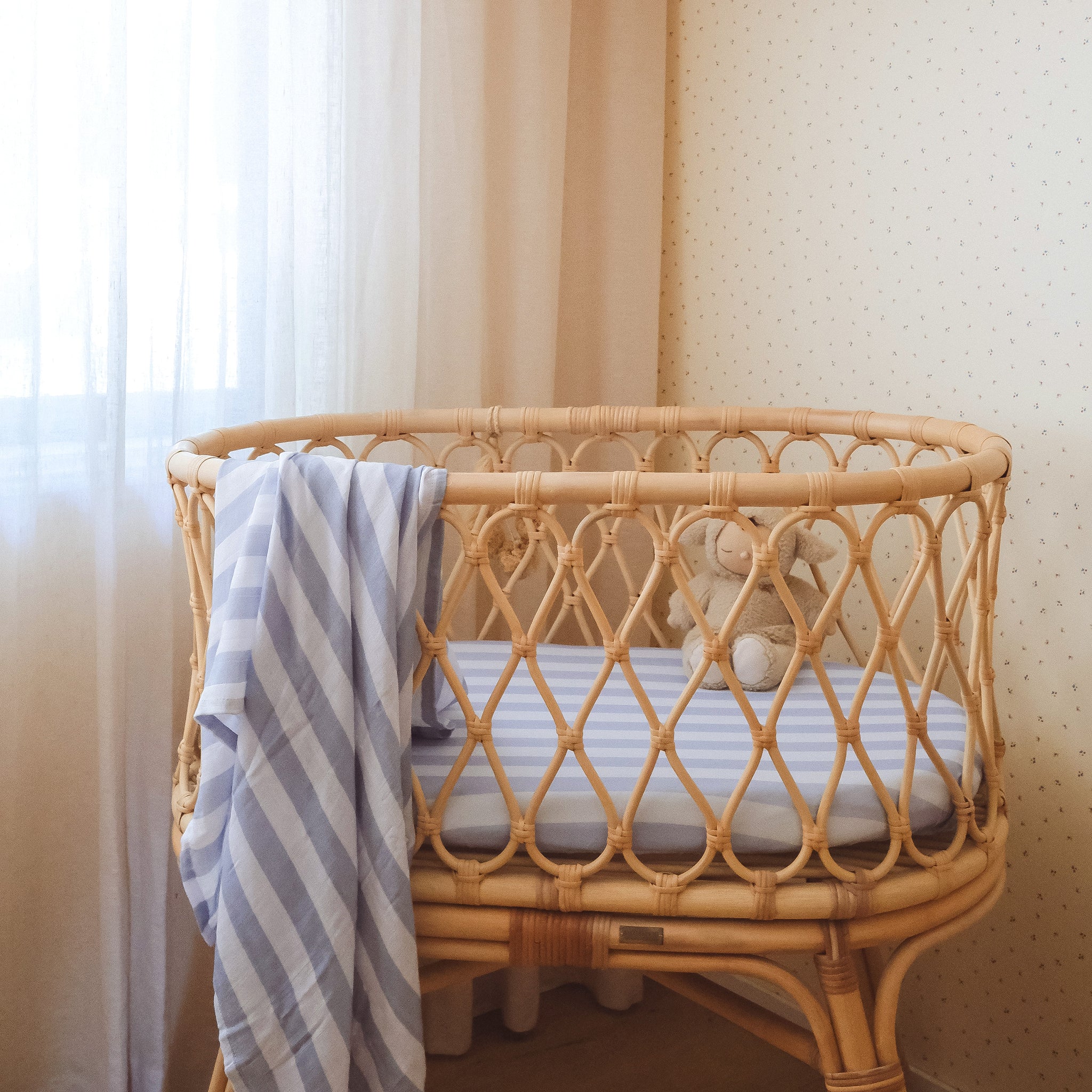 A cozy wicker crib features the FOX & FALLOW Cloud Stripe Organic Fitted Bassinet Sheet, a blue and white striped blanket, and a small beige teddy bear inside, set in a softly lit nursery with light curtains and dotted wallpaper.