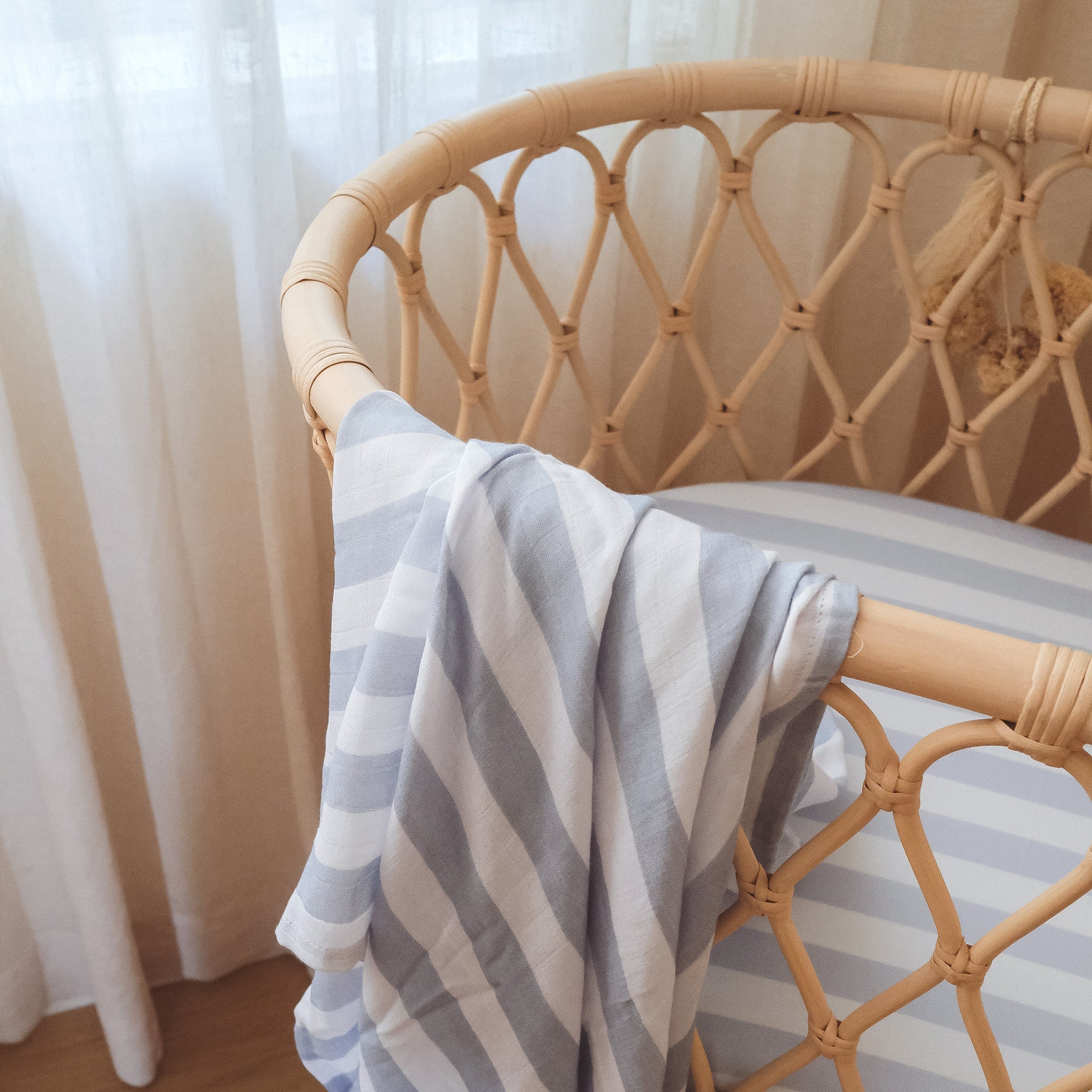 A rattan crib with a Fox & Fallow - Cloud Stripe Organic Fitted Bassinet Sheet/Change Pad Cover, made from GOTS certified material, sits by a window with sheer white curtains softly filtering natural light.
