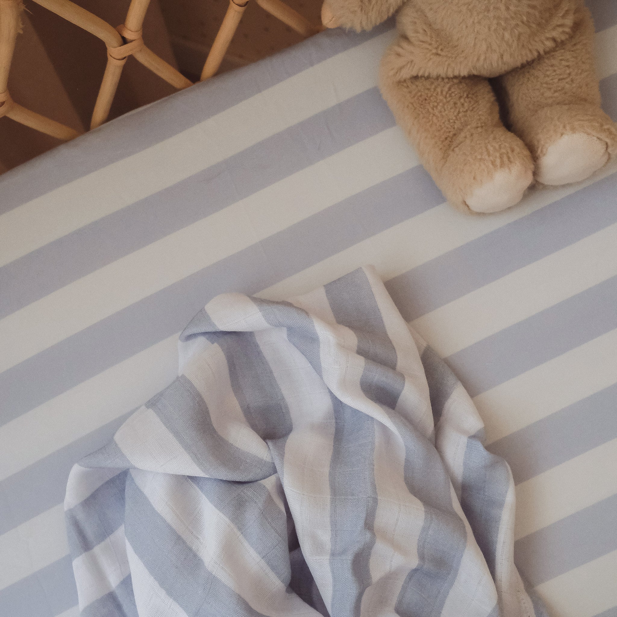 A close-up of the FOX & FALLOW Cloud Stripe Organic Fitted Bassinet Sheet/Change Pad Cover in blue and white stripes, styled with a matching blanket and a plush teddy bear in the crib corner.
