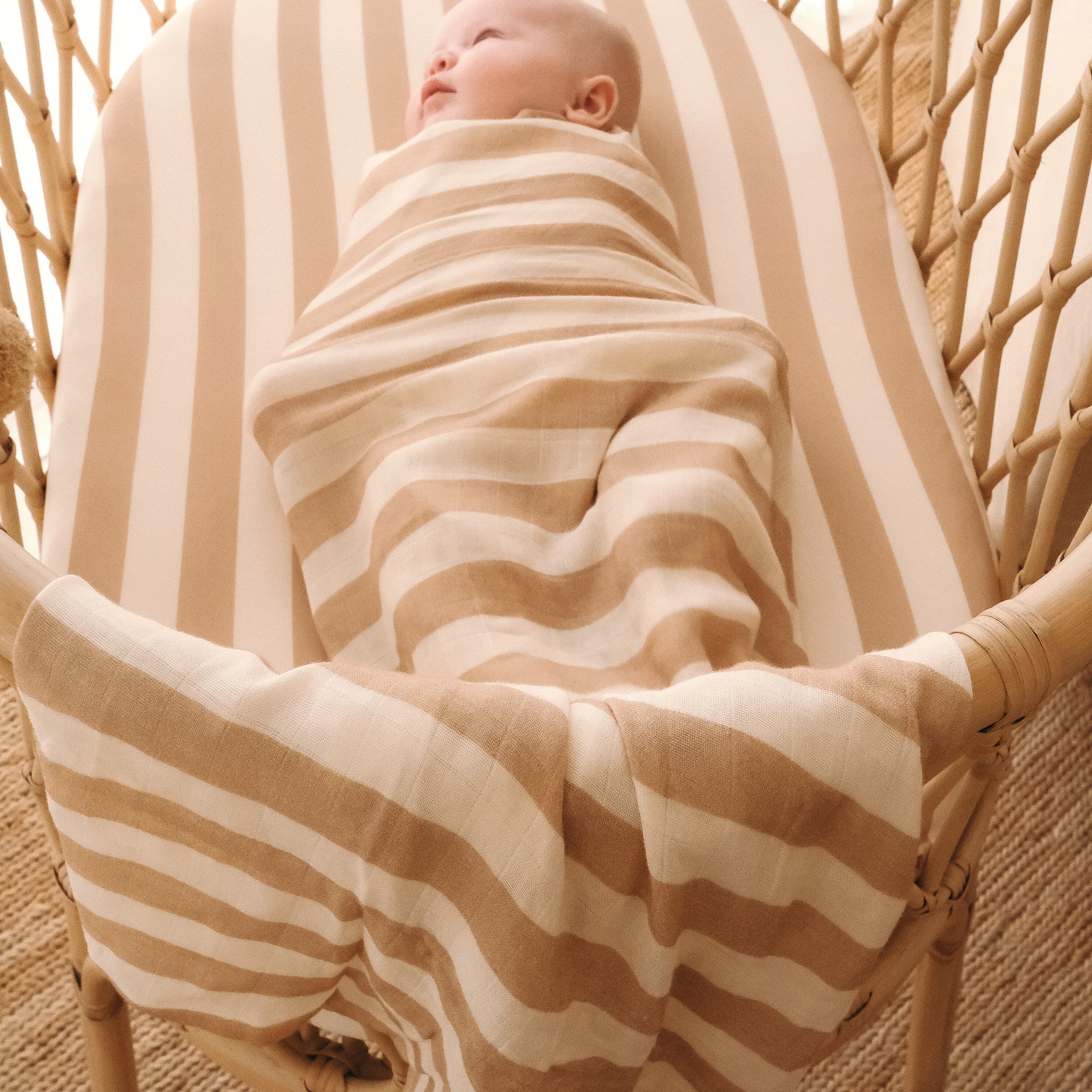 A baby rests in a rattan crib, lying on the FOX & FALLOW Coffee Stripe Organic Fitted Bassinet Sheet/Change Pad Cover. Cozy lighting and natural tones create a warm, inviting setting.