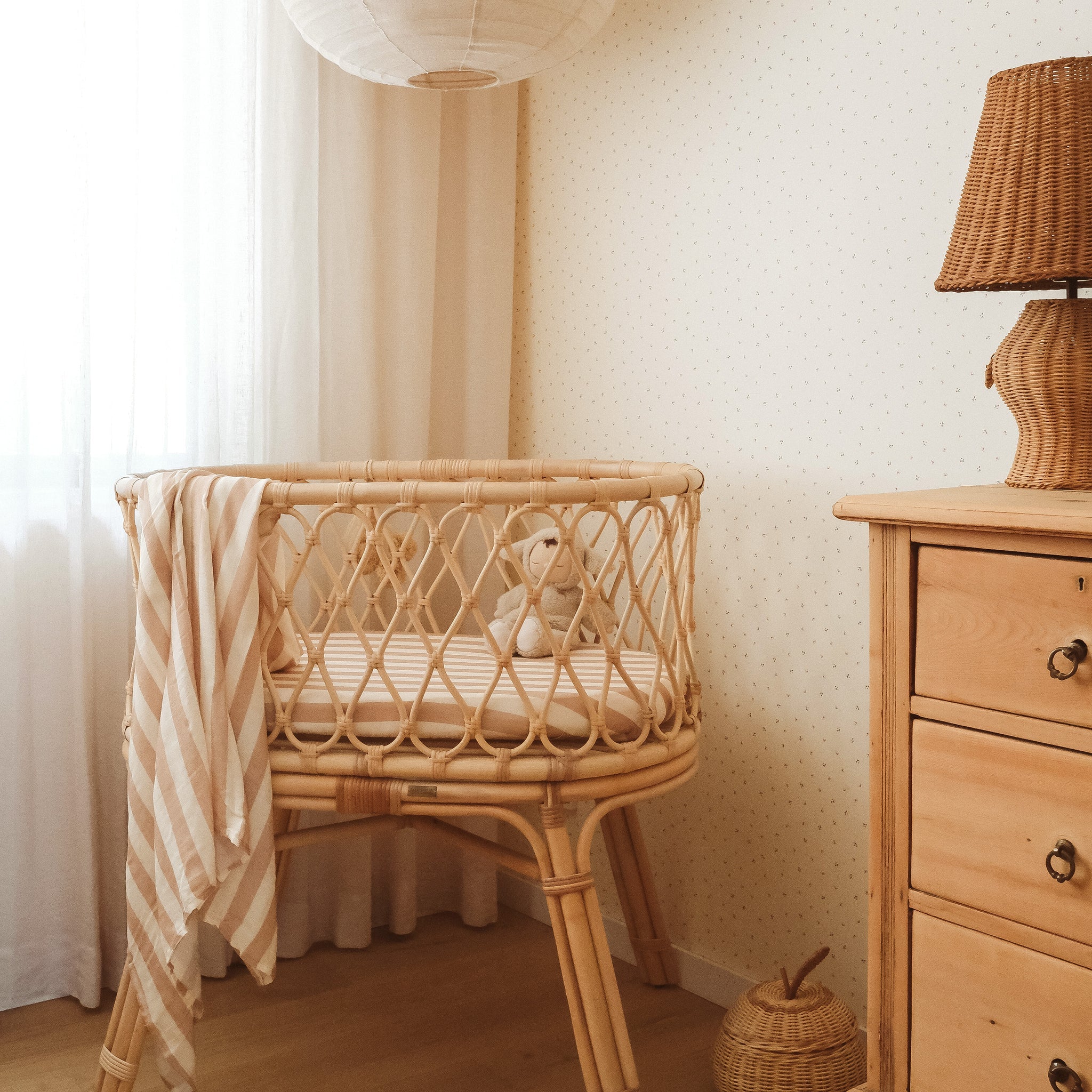 A cozy nursery with a wicker bassinet featuring the FOX & FALLOW Coffee Stripe Organic Fitted Bassinet Sheet/Change Pad Cover, holding a teddy bear and striped blanket. A wicker lamp and wooden dresser in soft neutral tones complete the scene with sheer curtains behind.