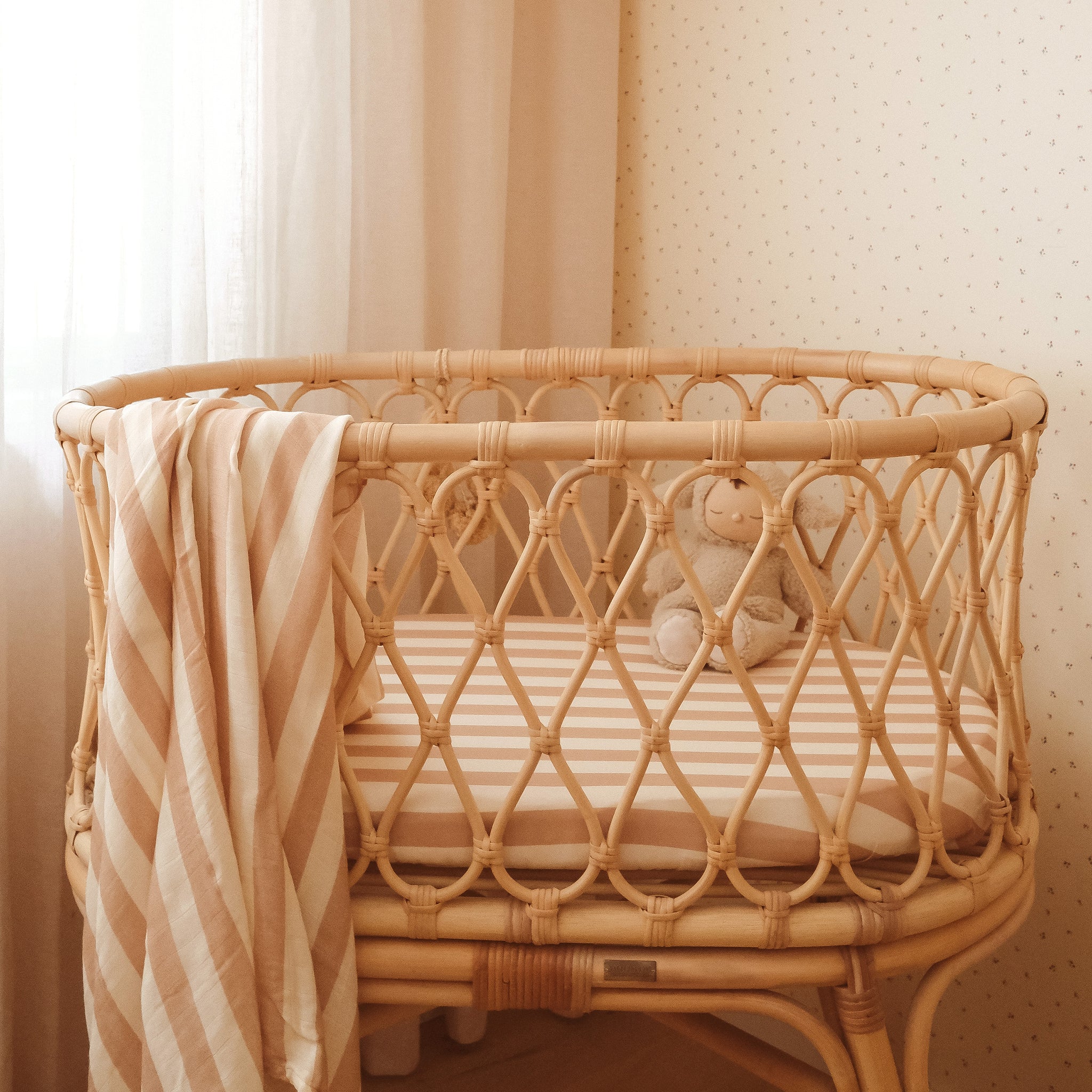 A woven rattan bassinet with the FOX & FALLOW Coffee Stripe Organic Fitted Bassinet Sheet, a beige and white blanket draped over the side, and a small stuffed animal sits against softly lit dotted wallpaper and a sheer curtain.