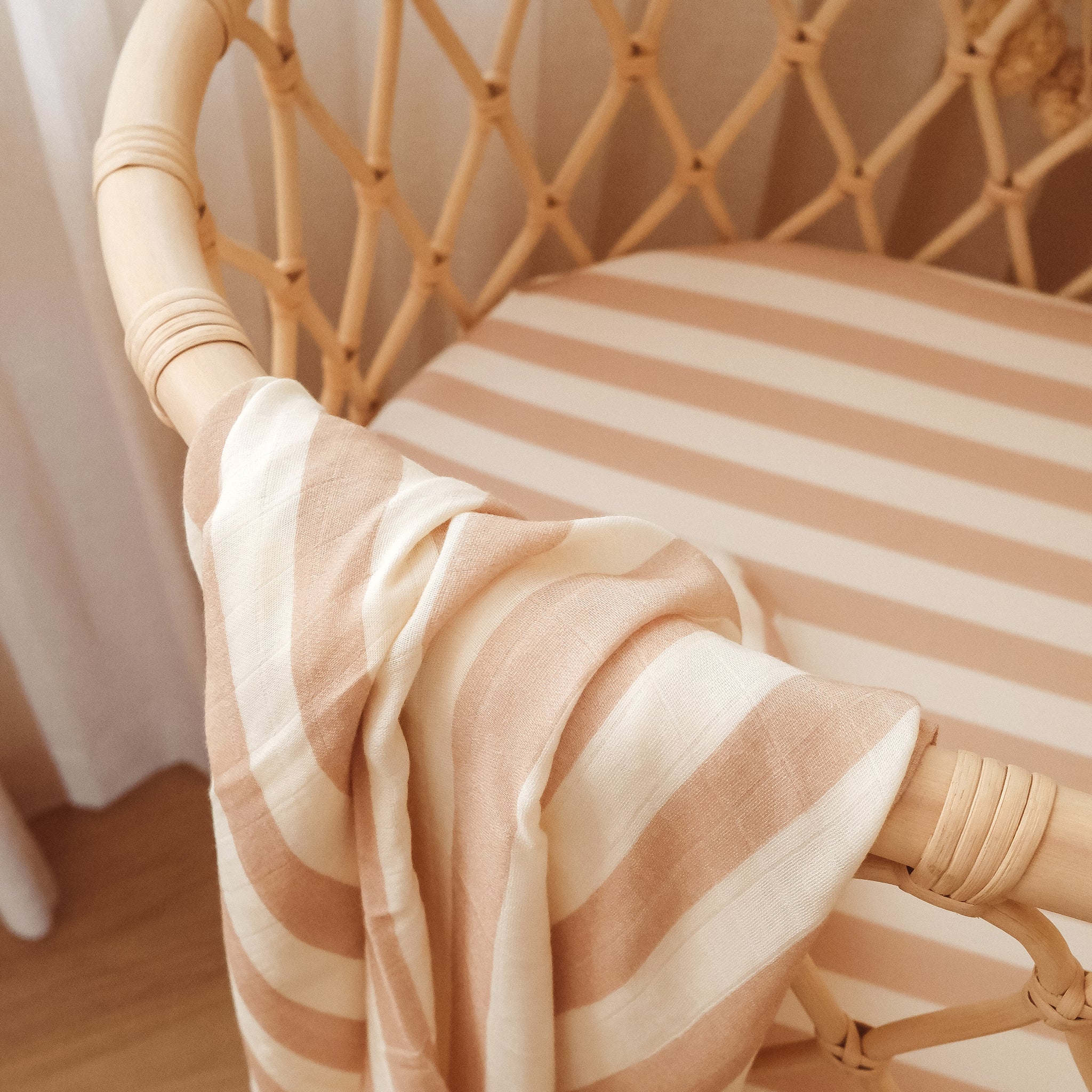 A rattan crib features the FOX & FALLOW Coffee Stripe Organic Fitted Bassinet Sheet/Change Pad Cover with a matching blanket, creating a cozy, softly lit, and inviting atmosphere for your baby.