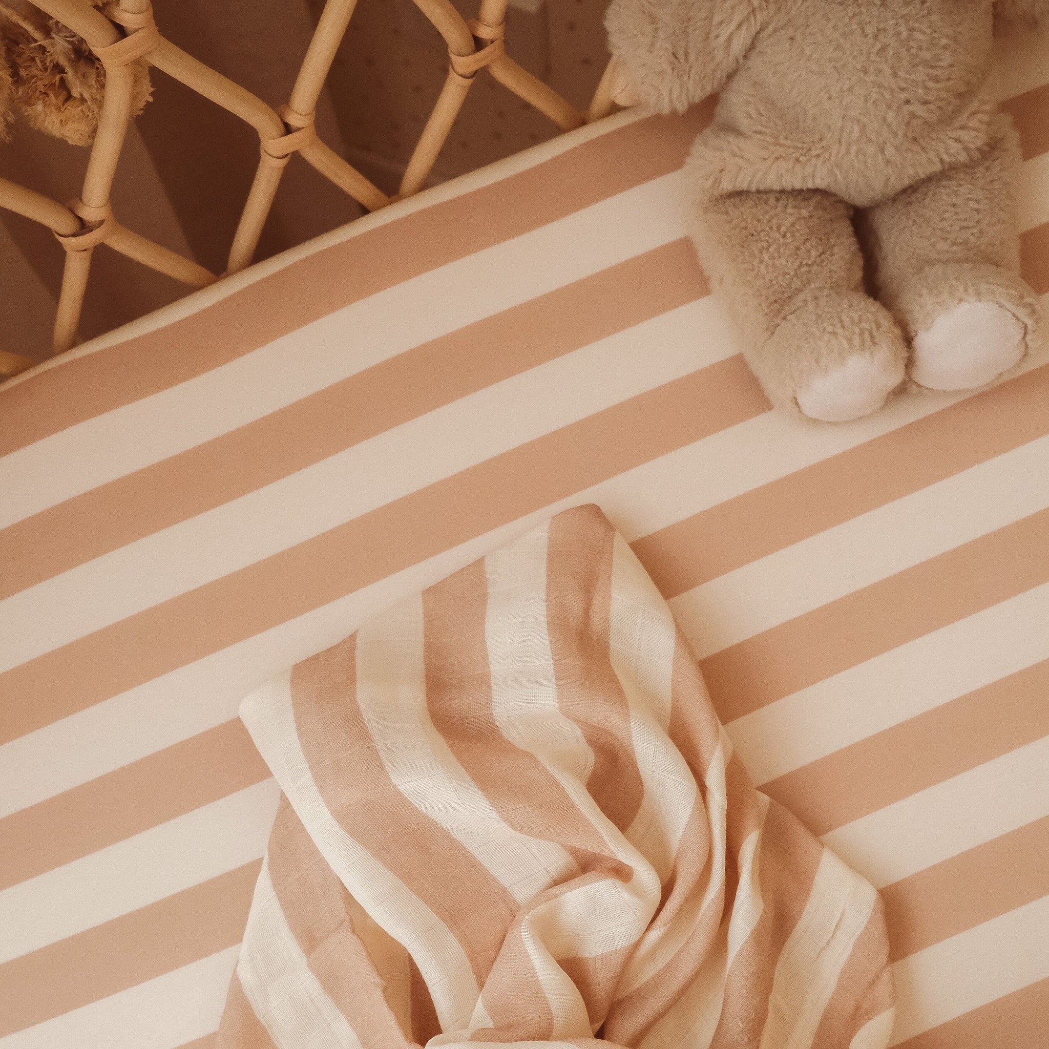 The FOX & FALLOW Coffee Stripe Organic Fitted Bassinet Sheet/Change Pad Cover, in soft beige and white stripes, adds comfort to the crib. GOTS certified baby bedding complements a plush toy near the wooden crib side.