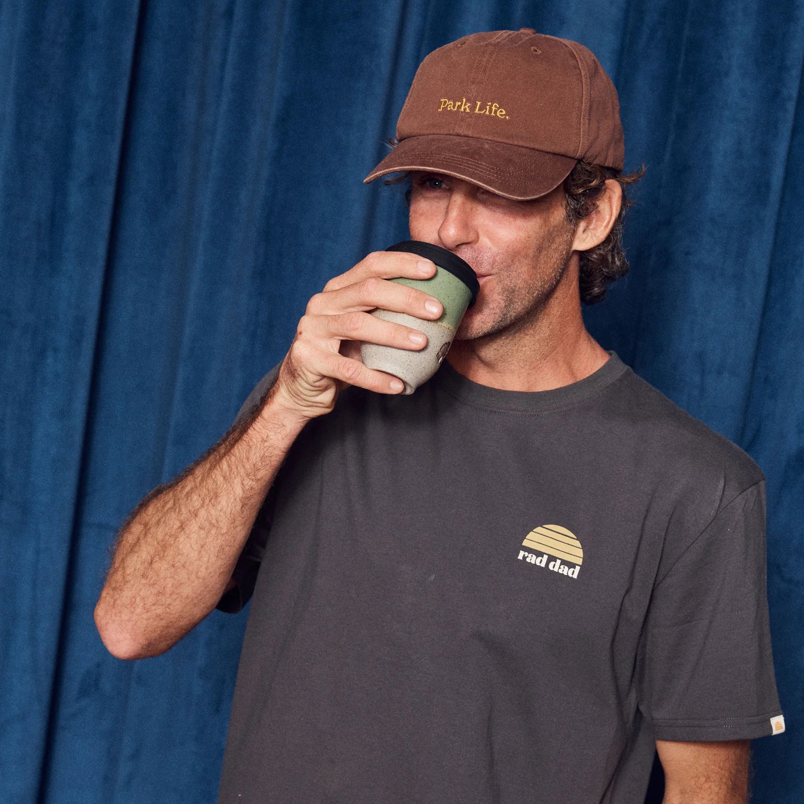 A man in a brown "Park Life." cap sips from a cup, wearing the BANABAE Banabae - Rad Dad Organic Cotton Tee Washed Black—perfect as a Dad gift or Father’s Day present—while standing in front of a blue curtain.