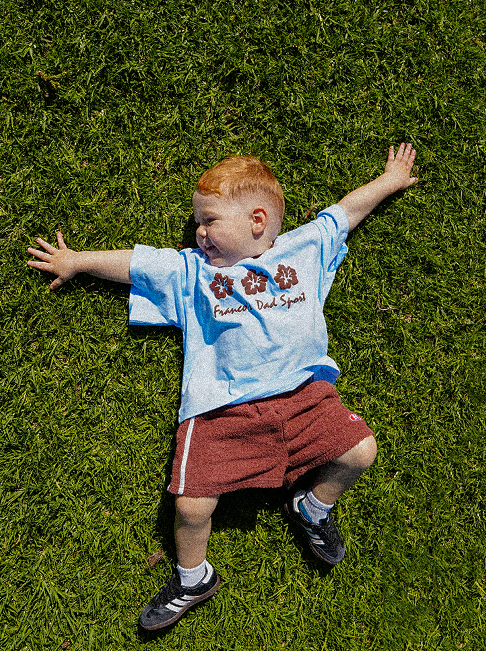 A young child with red hair is lying on a grassy field, wearing an oversized PRE-ORDER Hibiscus Tee from FRANCO'S DAD and red shorts, complemented by black and white sneakers. The child is spread-eagle, gazing to the side.