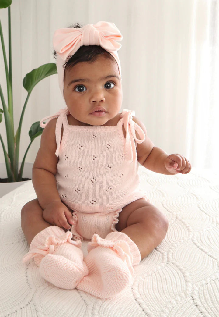 A baby wearing the Frill Bodysuit Primrose from ZIGGY LOU, complete with a headband featuring a large bow and matching knitted booties, is sitting on a white textured blanket. The 100% cotton ensemble captures the baby's curious expression as they look towards the camera. A green plant is visible in the background.