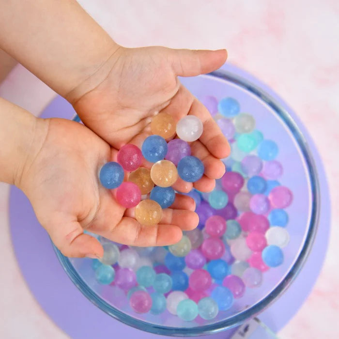 A child enjoys sensory play with several colorful Unicorn Bubbles Pastel Biodegradable Water Beads from NO NASTIES, holding them over a bowl filled with more. The blue, pink, purple, and clear beads ignite curiosity as they sit on a pastel purple surface, gently improving fine motor skills.