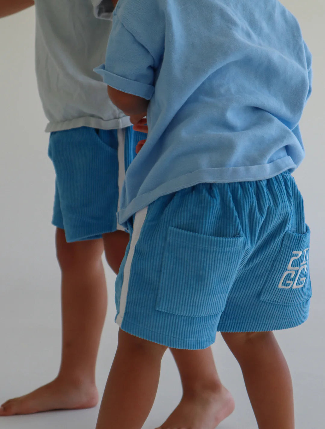 Two young children are dressed in light blue clothing, featuring "Cord Shorts River" with an elastic waistband and light blue shirts. They stand closely together, and one child has a number printed on the back pocket of their ZIGGY LOU shorts. The background is plain and light-colored.