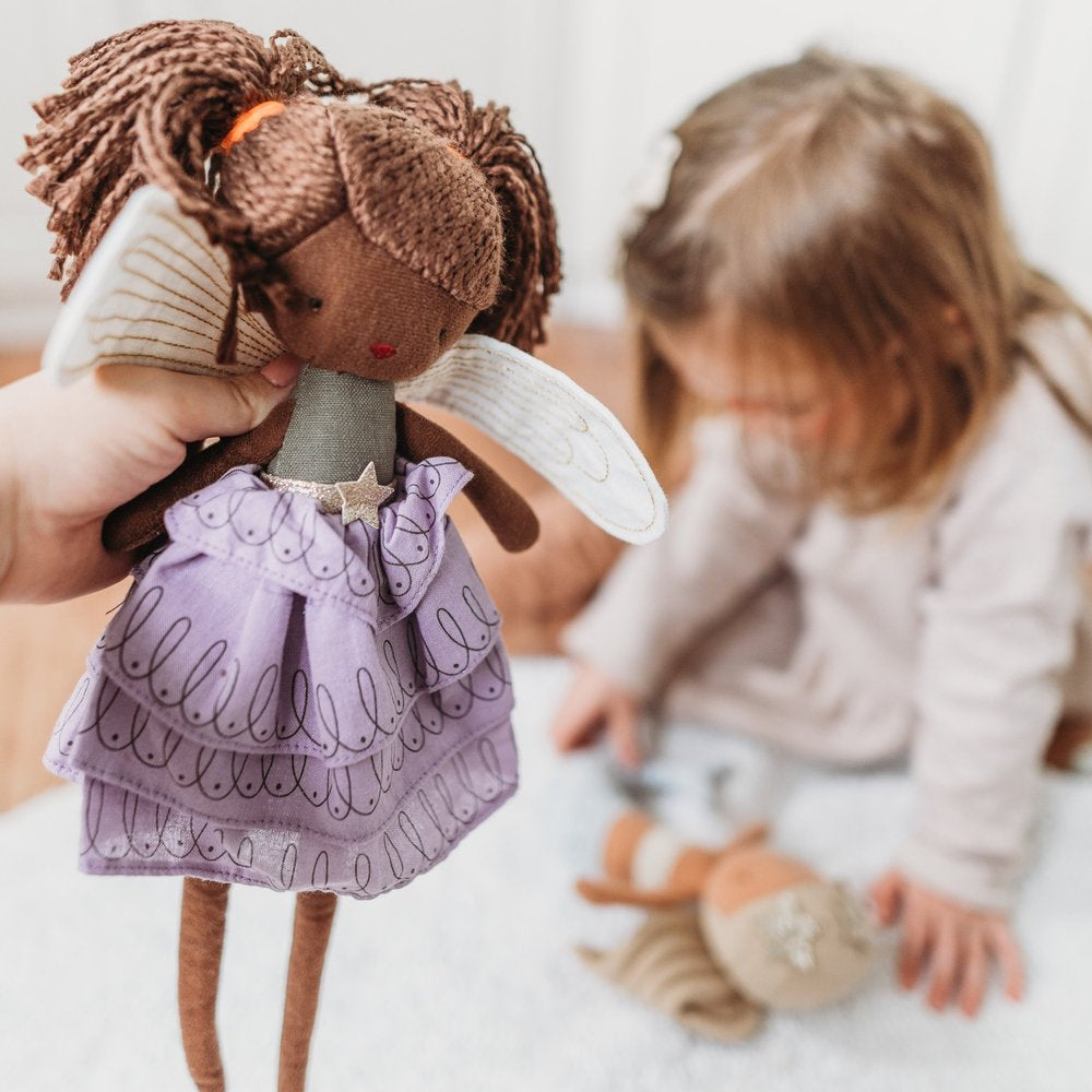 A child playing with a Bon Ton Toys brown-skinned doll with yarn hair and a shiny purple dress, while another Fairy Feline 35cm doll from PICCA LOULOU, complete with wings, is held up in the foreground. The background shows the child sitting on a white rug, focusing intently on the doll in their hands.