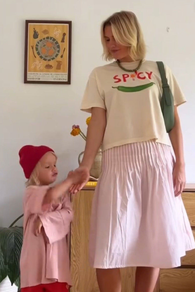 A woman wearing the MAKU FENAROLI Spicy T-Shirt, a beige boxy crop tee with "SPICY" emblazoned on it, pairs it with a pleated skirt as she stands in a room. Beside her, a child is dressed in a pink outfit and red beanie. They hold hands under a framed picture on the wall. The t-shirt showcases hand-painted designs and is made from 100% cotton.