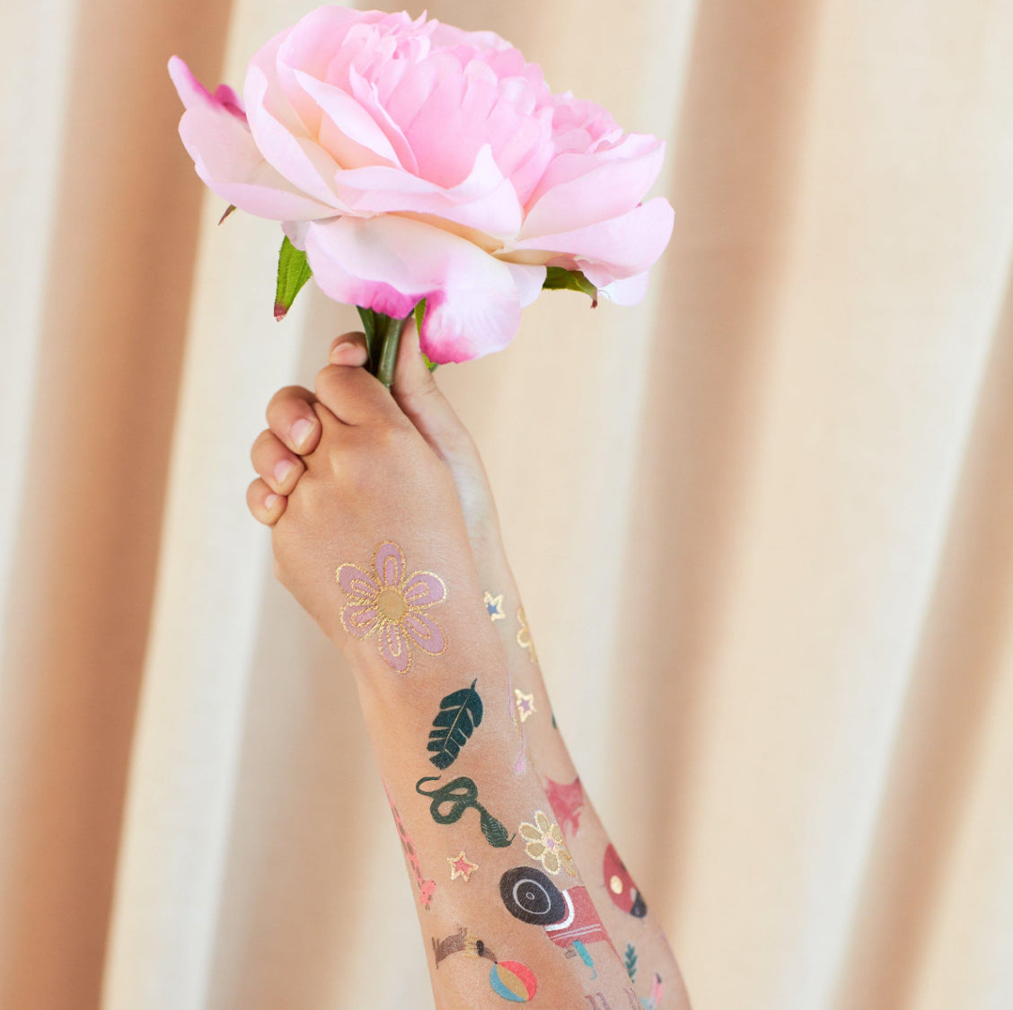 A child's hand holding a large, light pink rose against a beige curtain background. The arm is adorned with various colorful temporary tattoos from the MERI MERI Fairy Tattoos (Pack of 2), featuring shapes like flowers, leaves, stars, and infinity symbols. These whimsical designs are reminiscent of a fairy party setting and include hints of shimmering gold foil accents.