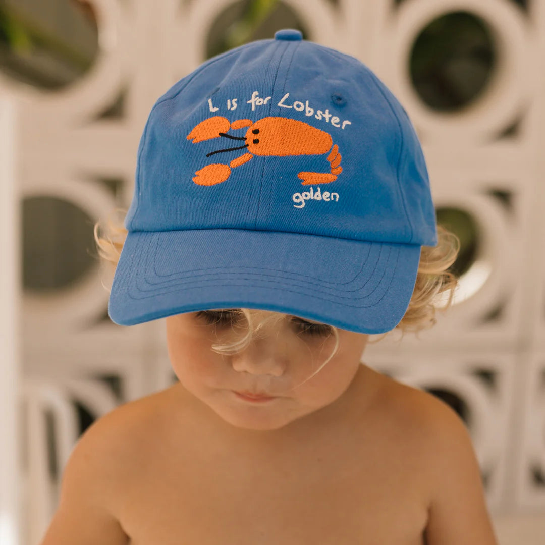 A child wearing the GOLDEN CHILDREN Lobster Cap in Deep Sea Blue, which features an embroidered orange lobster and the text "L is for Lobster golden." An adjustable brass clasp adds a touch of detail. The child looks down with a neutral expression, set against a softly blurred background.