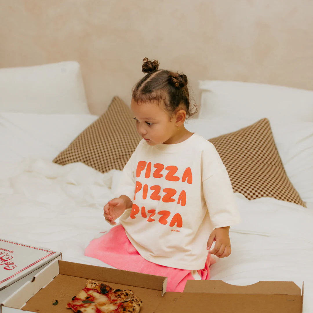 A young child with hair in two buns sits on a bed wearing the soft stretch Golden Children ~ Pizza Mid Sleeve Tee Eggnog. "Pizza" is written in orange, matching the pizza boxes nearby. Cozy checkered pillows and a neutral wall complete the scene.