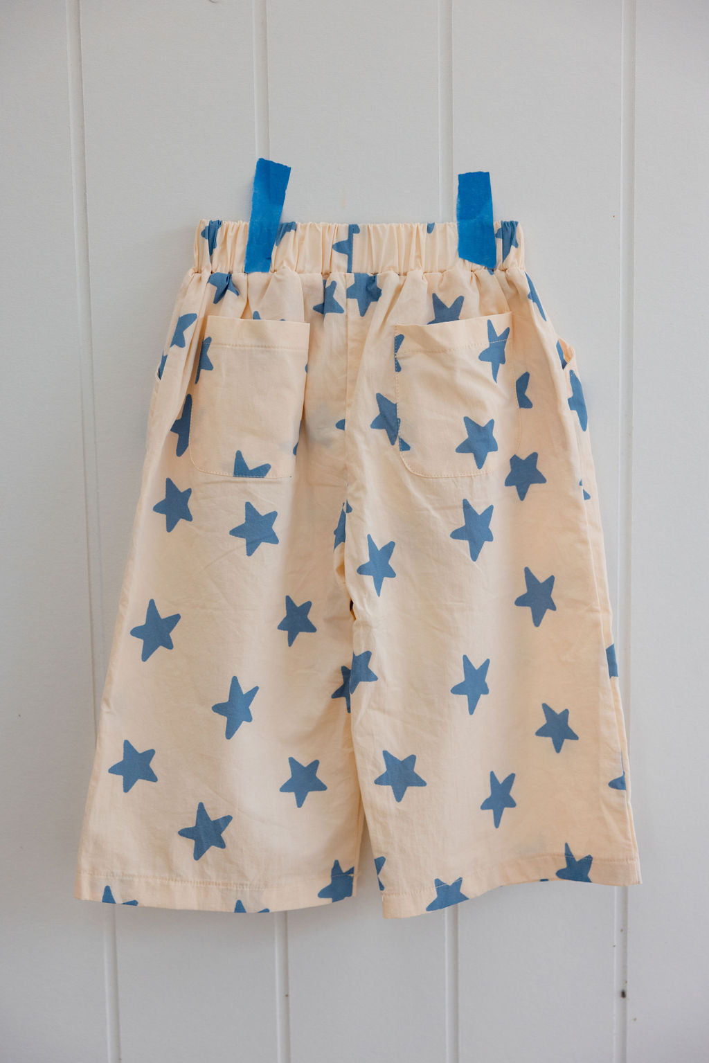 The Sunday Siblings ~ Dusty Star Pants by SUNDAY SIBLINGS are beige cotton shorts with blue star patterns, featuring an elastic waistband and two back pockets, stylishly displayed on a wall secured by blue tape.