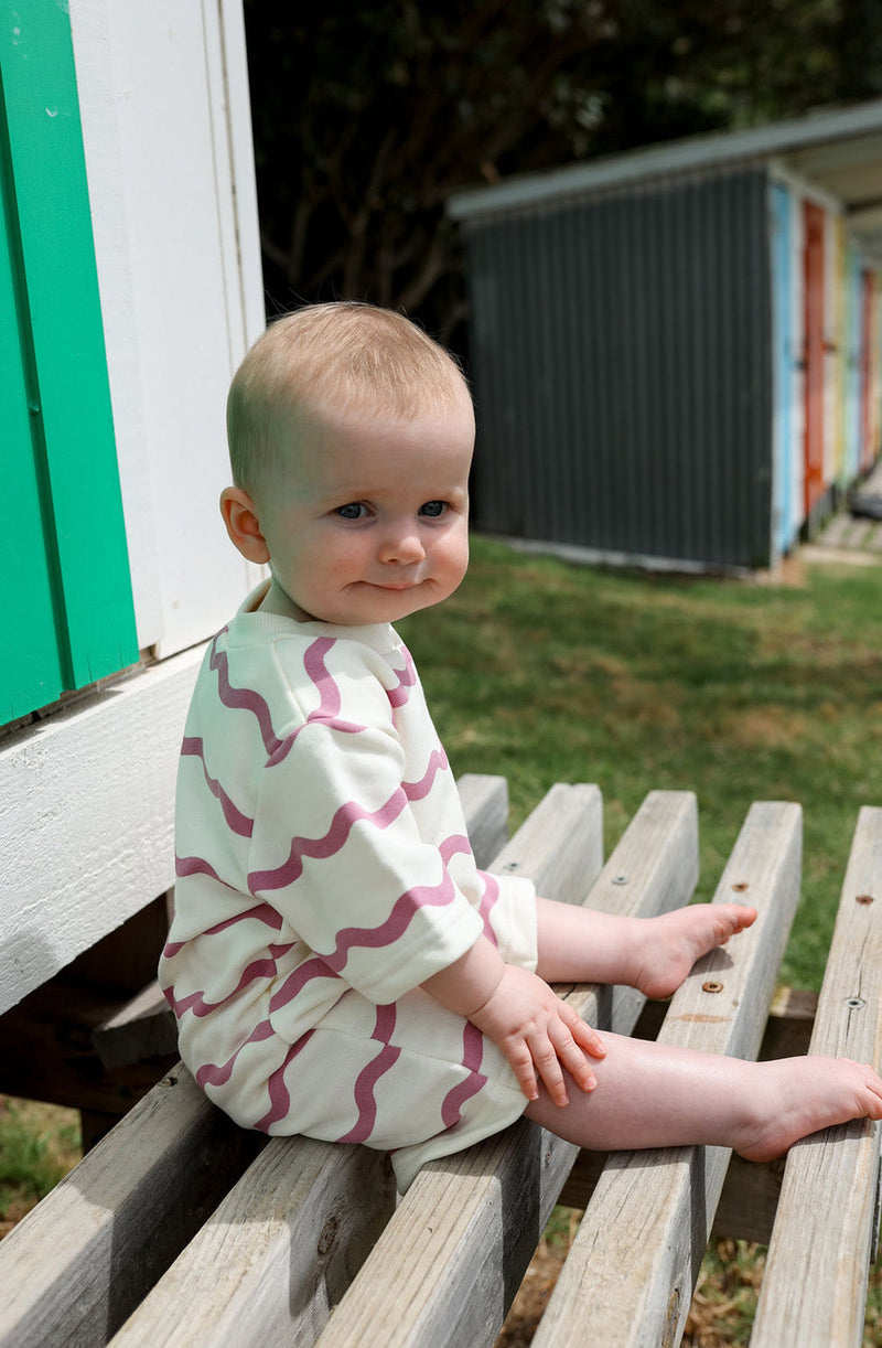 Baby sitting on timber chair wearing the Bebe romper in rosa wiggle print