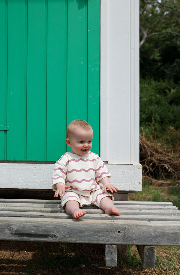 Baby sitting against green and white shed wearing the romper in rosa wiggle print