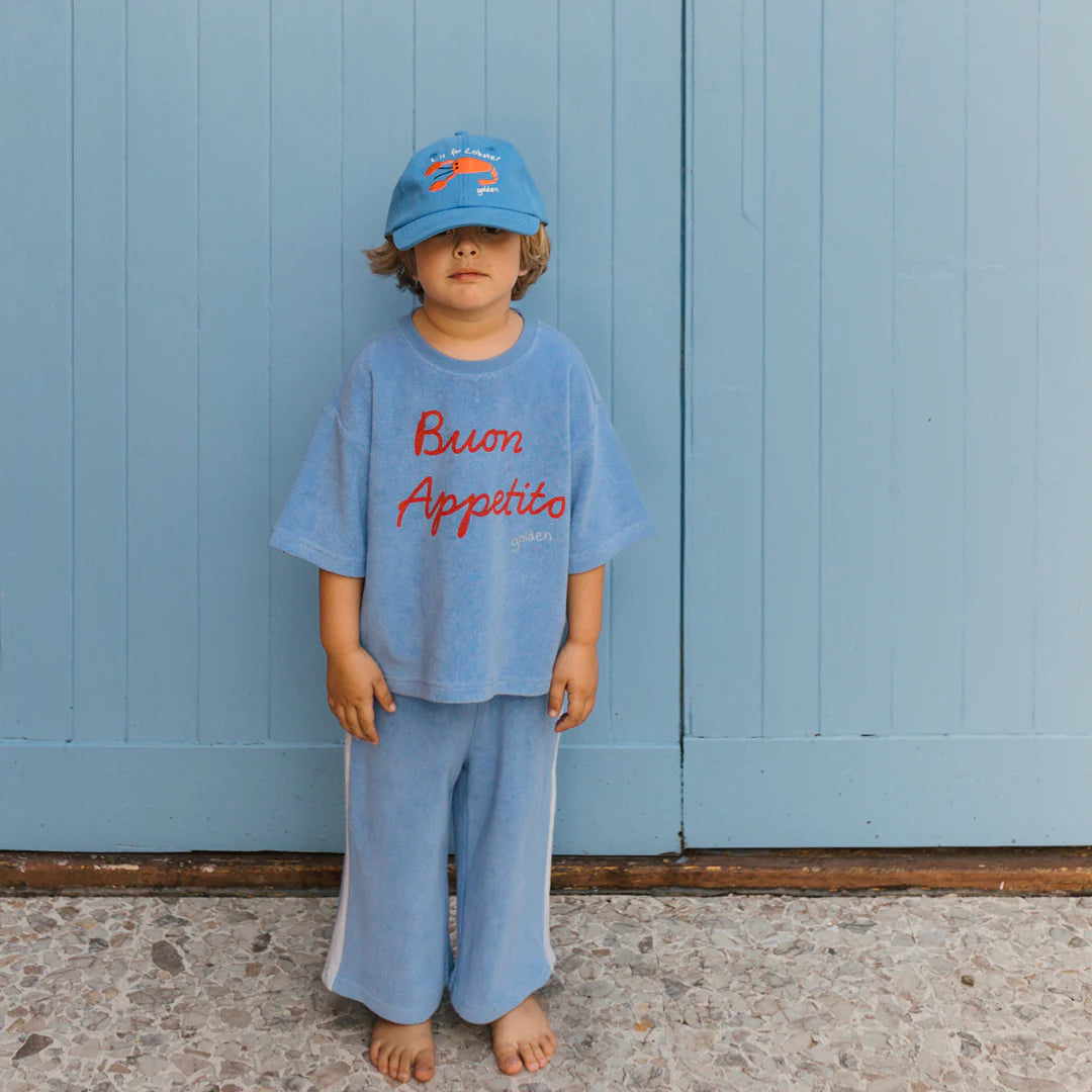 A young child stands barefoot against a light blue wall, dressed in a blue outfit and an adorable Lobster Cap Deep Sea Blue from GOLDEN CHILDREN, featuring an adjustable brass clasp. The shirt displays "Buon Appetito" in red, adding to the casual and relaxed vibe.