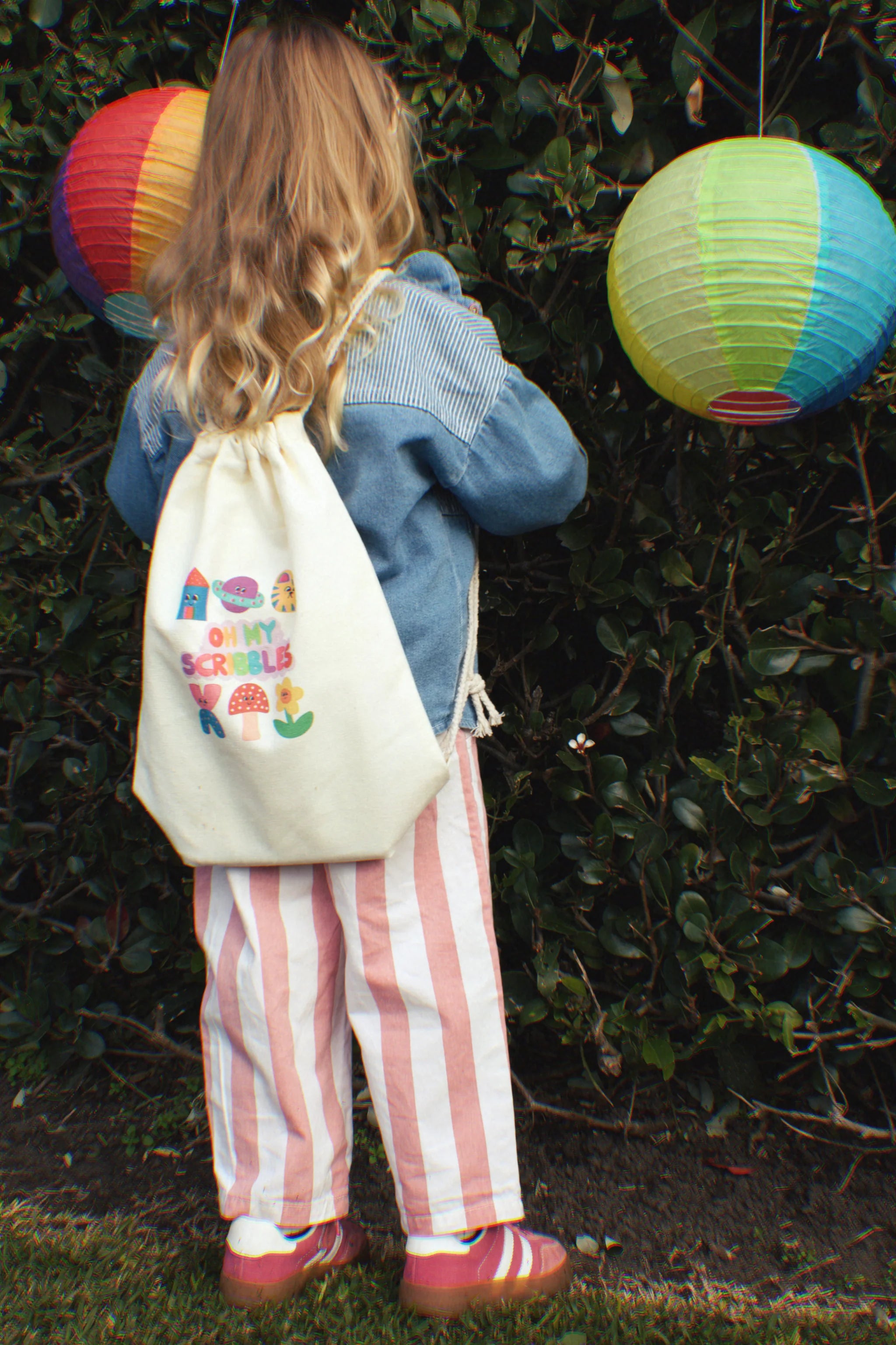 A child with long hair, wearing a denim jacket and pink-and-white striped pants, stands with their back to the camera. They hold an "Oh My Scribbles Reusable Colouring Book" by OH MY FRANKIE, ready to unleash creativity near a hedge adorned with colorful paper lanterns.