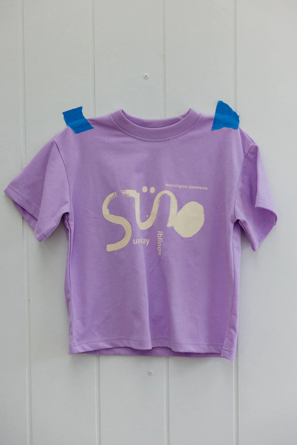 The Sunday Siblings Il Sole Tee in purple/cream, known for its oversized fit and soft cotton fabric, is taped to a light-colored wall using blue tape. It features graphic designs and the words "mornington peninsula" and "sunlay.