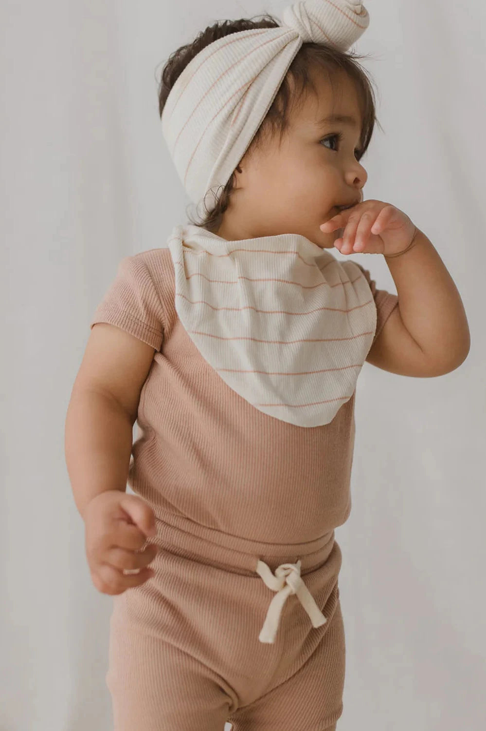 A toddler stands wearing a light pink ribbed outfit with a tie at the waist, accessorized with an organically made SUSUKOSHI Bandana Bib Coco Stripe—white with thin red stripes—and a matching headband with a bow. With one hand near their mouth and gazing to the side, the toddler is set against a plain, neutral background.