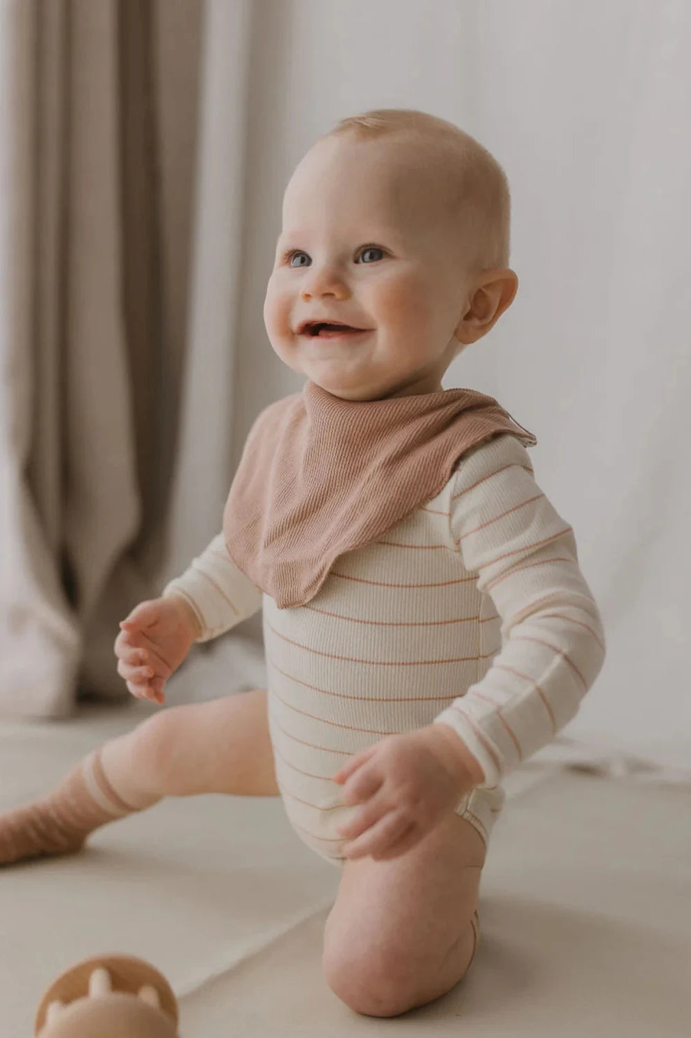 A smiling baby with short blond hair, dressed in a long-sleeve striped onesie and an ethically made organic cotton SUSUKOSHI Bandana Bib Terracotta, is kneeling on a light-colored floor. The background features soft fabric drapes in muted tones. The baby looks happy and engaged with their surroundings.