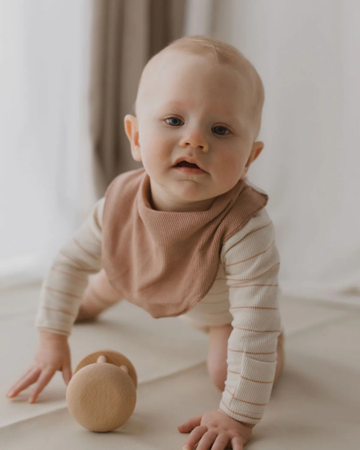 A baby with light hair, wearing a striped long-sleeved onesie and an ethically made Bandana Bib Terracotta by SUSUKOSHI, is crawling on a light-colored surface. A wooden rattle is placed in front of the baby. The background features soft, neutral-toned curtains.