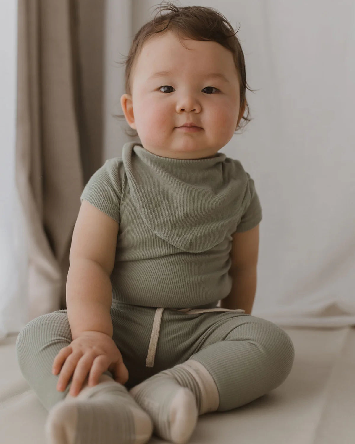 A baby sits on a light-colored surface against a neutral backdrop, wearing an ethically made, green ribbed outfit with matching leggings and socks. The baby has short, dark hair and is looking directly at the camera with a calm expression. A Bandana Bib Sage from SUSUKOSHI, made from organic cotton, completes the adorable look.