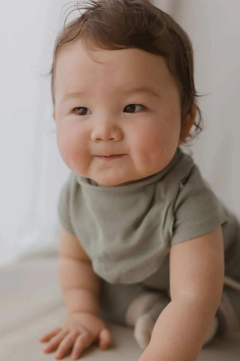 A baby with short, brown hair and chubby cheeks is shown crawling, wearing a light green shirt made of ethically produced organic cotton. The baby has a slight smile on their face, and the Bandana Bib Sage by SUSUKOSHI peeks out from under the shirt. The softly blurred background in neutral colors highlights the baby's expressive features.