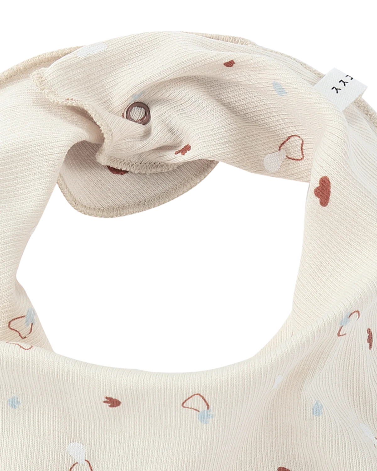 A close-up image of the Bandana Bib Sky by SUSUKOSHI, made from soft, textured organic cotton in beige. The bib showcases an adorable pattern of small hearts in muted shades of brown and blue. A tag visible on the upper right side highlights its ethical production with two printed letters.