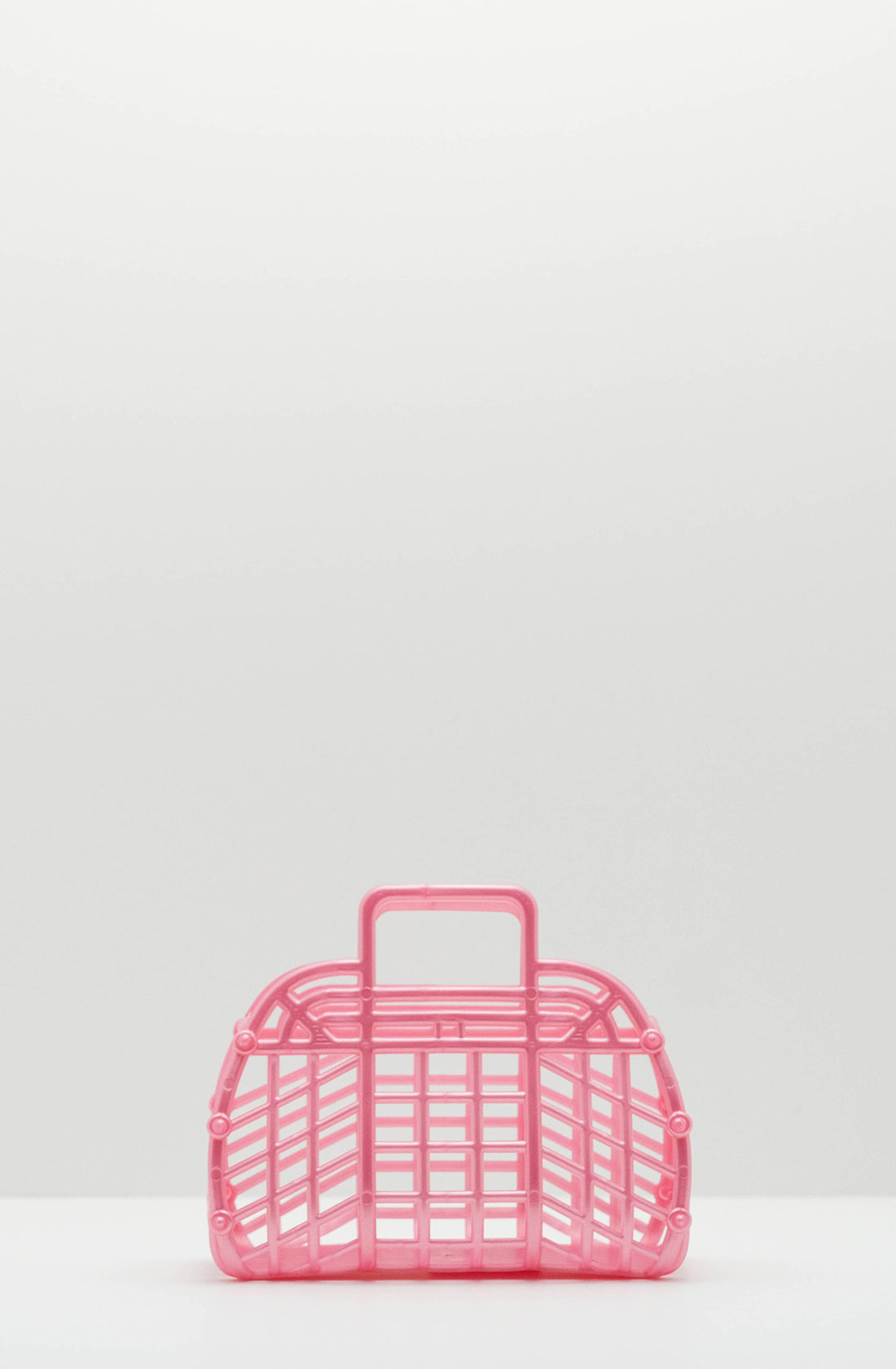 A Mini Retro Jelly Basket Pearl Pink by BAY KIDS, crafted from recyclable LDPE, sits on a plain, light-colored surface against a white background. This BPA-free handbag boasts a basket-like structure with a latticed design and features a handle on top.