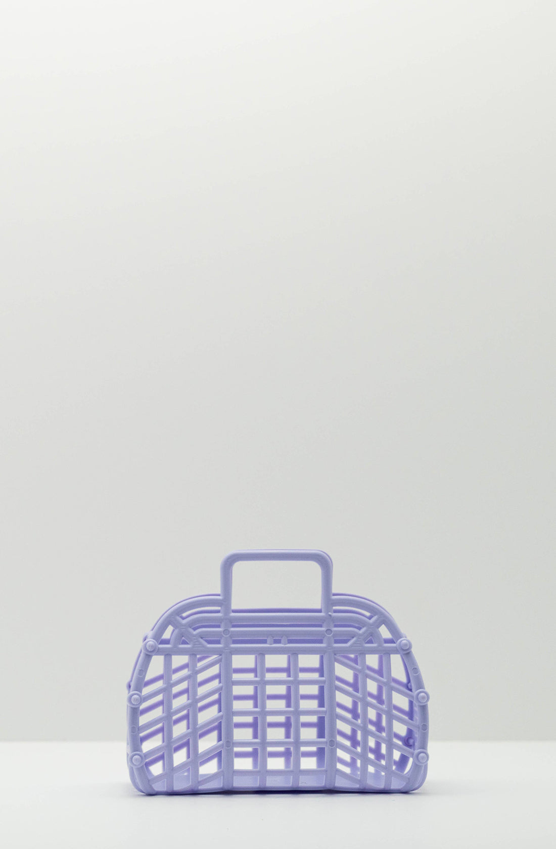 A Mini Retro Jelly Basket Lilac by BAY KIDS, crafted from recyclable BPA-free LDPE, stands on a white surface against a plain light gray background. This small lavender-colored plastic basket features an open grid design with a rectangular flat bottom, curved top, and single handle.