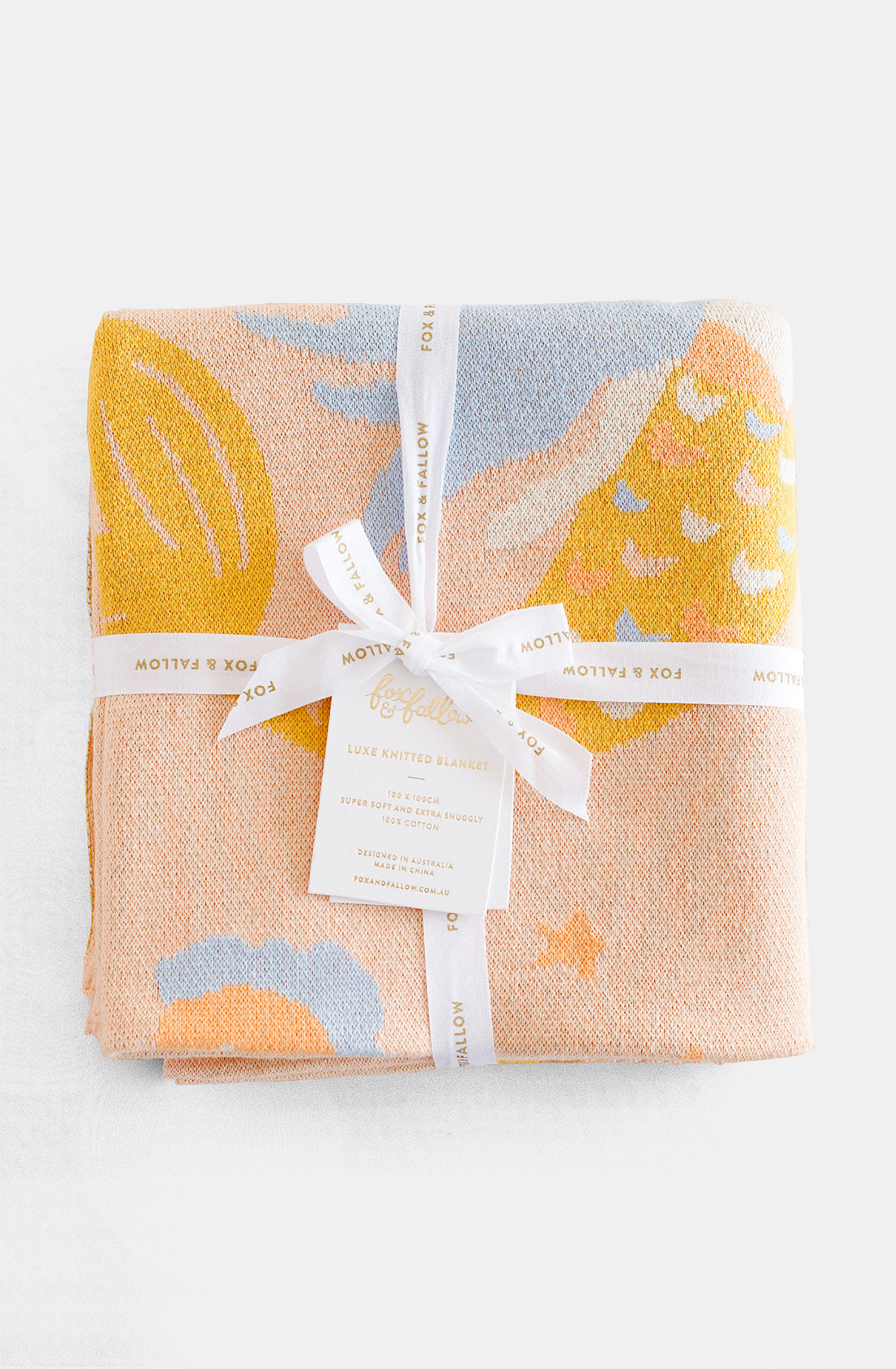 A neatly folded, pastel-colored Mermaids Blanket wrapped with a white ribbon adorned with the words "FOX & FALLOW." Crafted from 100% cotton, this luxe baby gift features whimsical patterns in tones of blue, yellow, and peach, with a label in the center detailing its luxury quality and brand name.