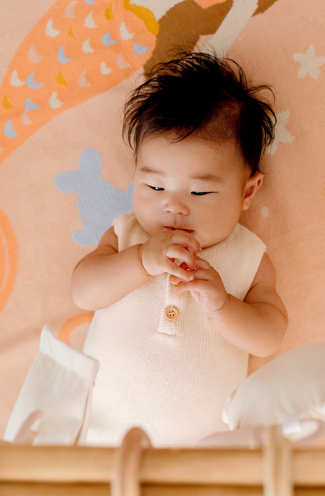 A baby with dark hair lies on the Mermaids Blanket by FOX & FALLOW, a luxe knitted blanket featuring peach hues, abstract shapes, and small fish designs. The baby is wearing a sleeveless, cream-colored onesie and is gazing downward while touching their hands together, with a soft focus on their playful expression—perfect for a thoughtful baby gift.