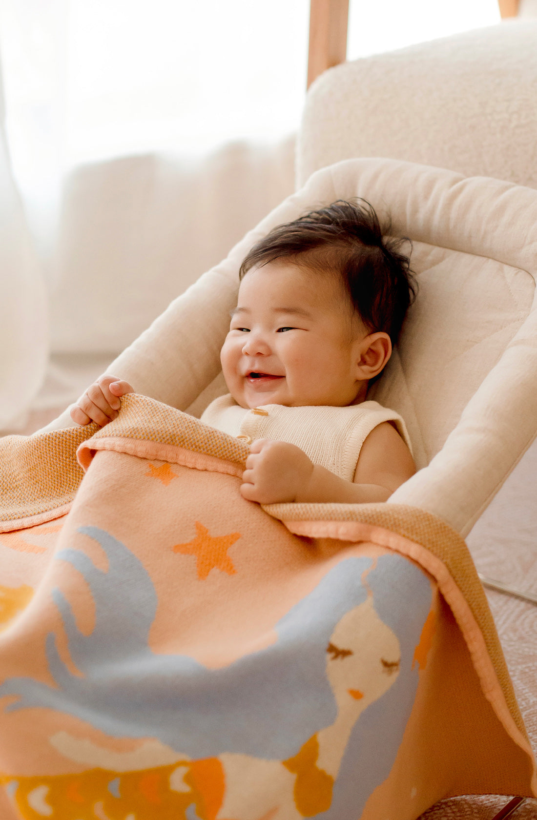 A smiling baby is sitting in a cushioned baby chair with a soft, colorful Mermaids Blanket from FOX & FALLOW, showcasing delightful starfish and fish designs. This stunning baby gift, made from 100% cotton, creates a cozy atmosphere. The baby's eyes are slightly closed as they enjoy the warm ambiance of the softly lit background.