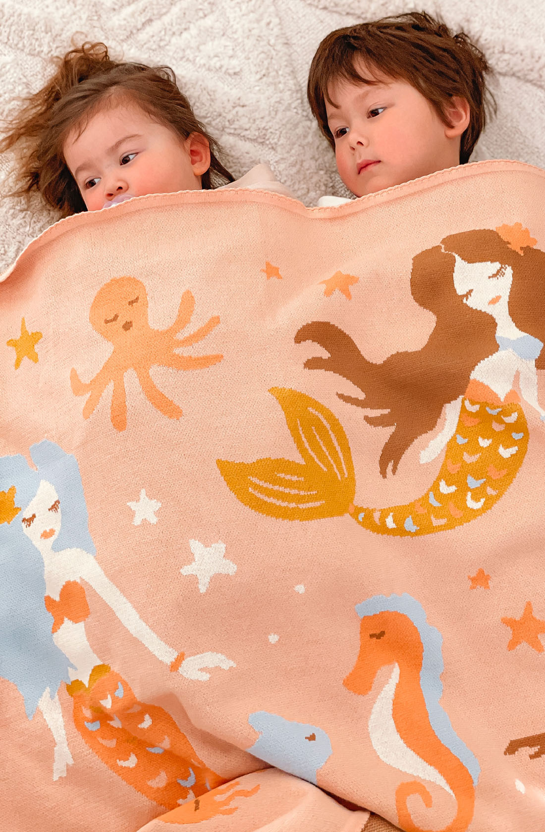 Two young children lie side by side under the FOX & FALLOW Mermaids Blanket, a peach-colored 100% cotton blanket adorned with whimsical sea-themed illustrations, including mermaids, an octopus, and seahorses. This luxe knitted blanket makes them appear calm and cozy as they gaze slightly off-camera—an ideal baby gift.