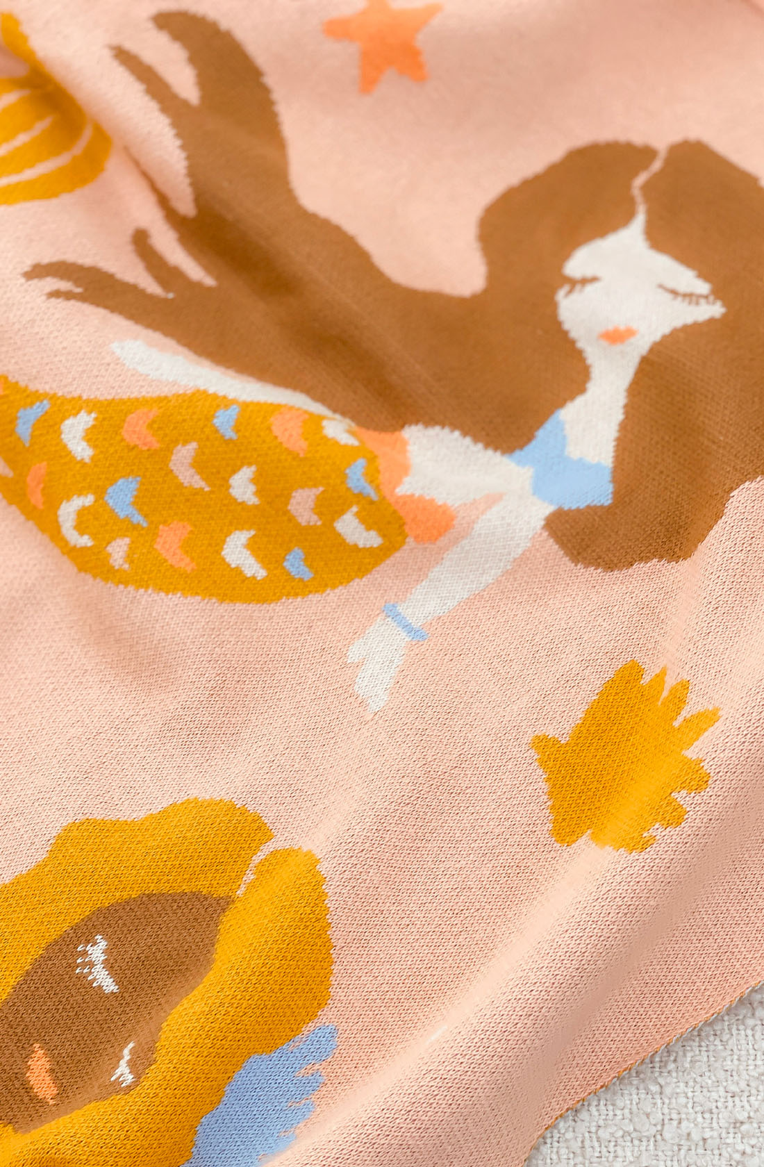 A close-up of the Mermaids Blanket by FOX & FALLOW, crafted from 100% cotton fabric. It features a whimsical design of a mermaid with flowing brown hair, an orange fish tail adorned with colorful scales, and a blue top. The soft peach background is accented with decorative elements like yellow seaweed and starfish—making it perfect for luxe knitted blankets as a baby gift.