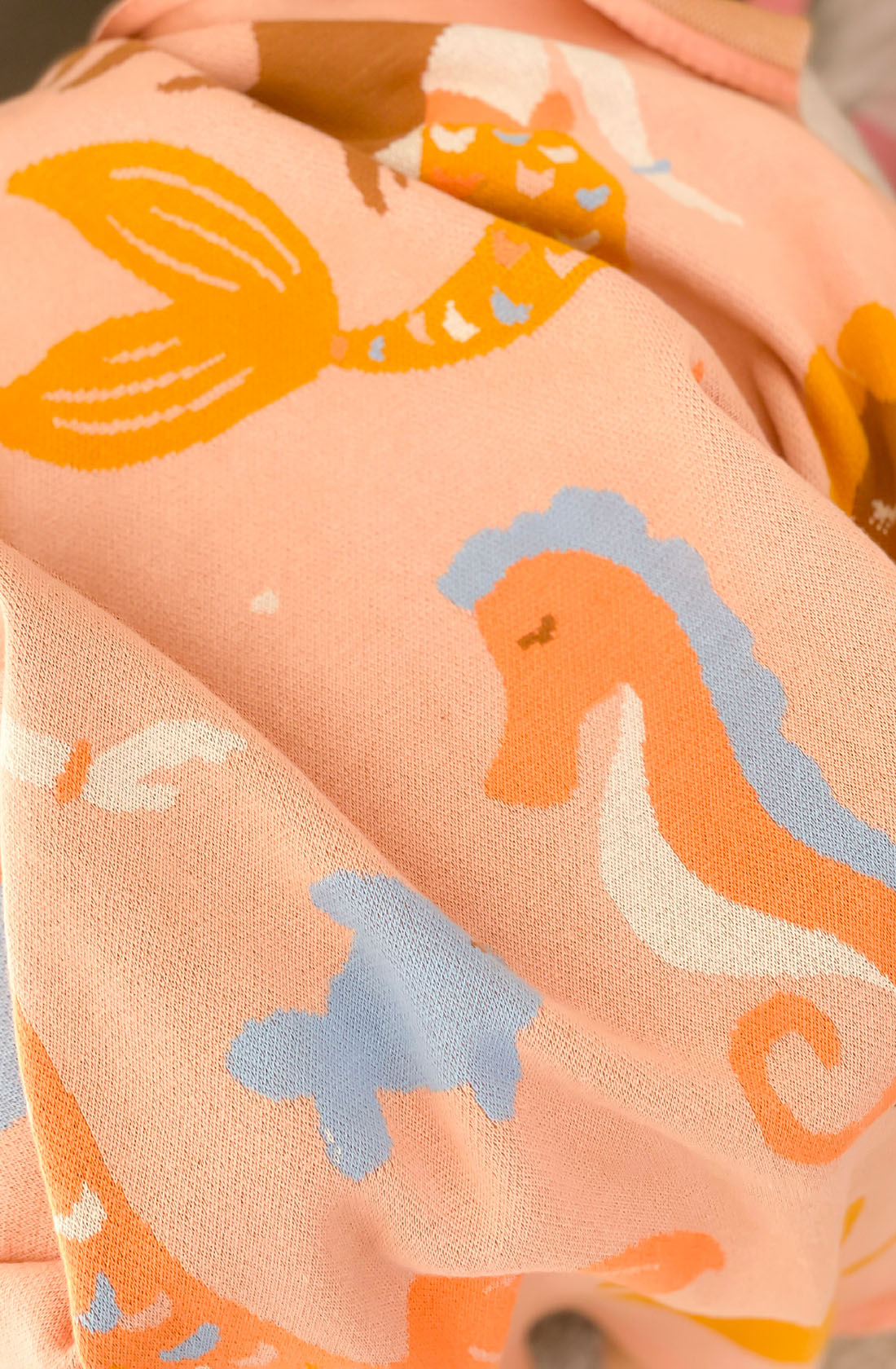 A detailed view of the Mermaids Blanket by FOX & FALLOW, made from 100% cotton fabric in a delightful peach hue. The aquatic-themed design showcases orange mermaids, orange and blue seahorses, and blue sea turtles. The fabric looks soft with visible folds and textures—perfect for crafting luxe knitted blankets or as a considerate baby gift.