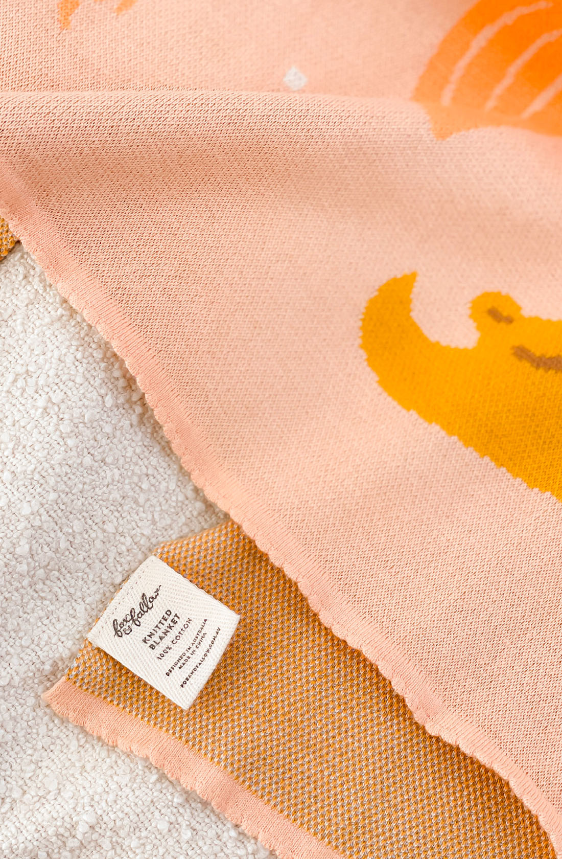 A close-up of the Mermaids Blanket by FOX & FALLOW, featuring a knitted peach-colored design with an orange pattern, draped over a white textured surface. A small fabric tag reading "forcalor" is visible near the bottom left corner of this luxurious blanket made from 100% cotton—ideal as a baby gift.