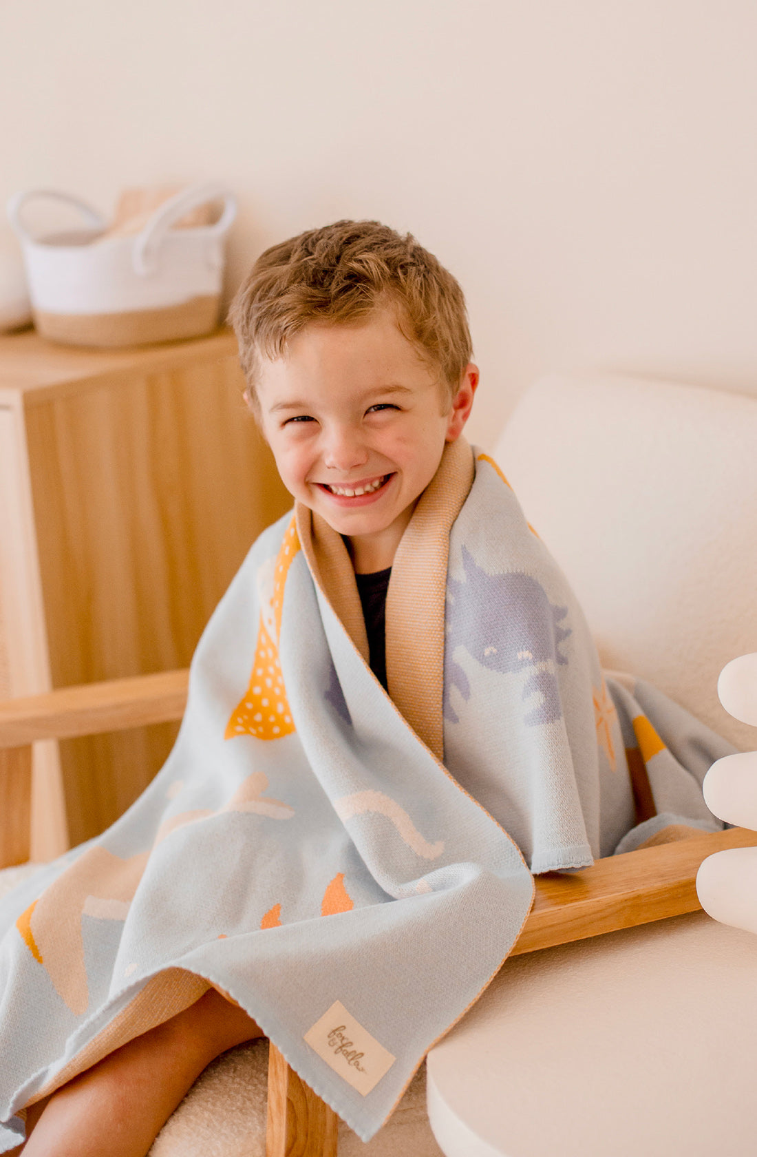 A young boy is smiling while sitting on a wooden chair. He is wrapped in the Ocean Creatures Blanket by FOX & FALLOW, a light blue 100% cotton baby blanket adorned with playful animal illustrations. The room has a minimalist decor with wooden furniture and soft colors.