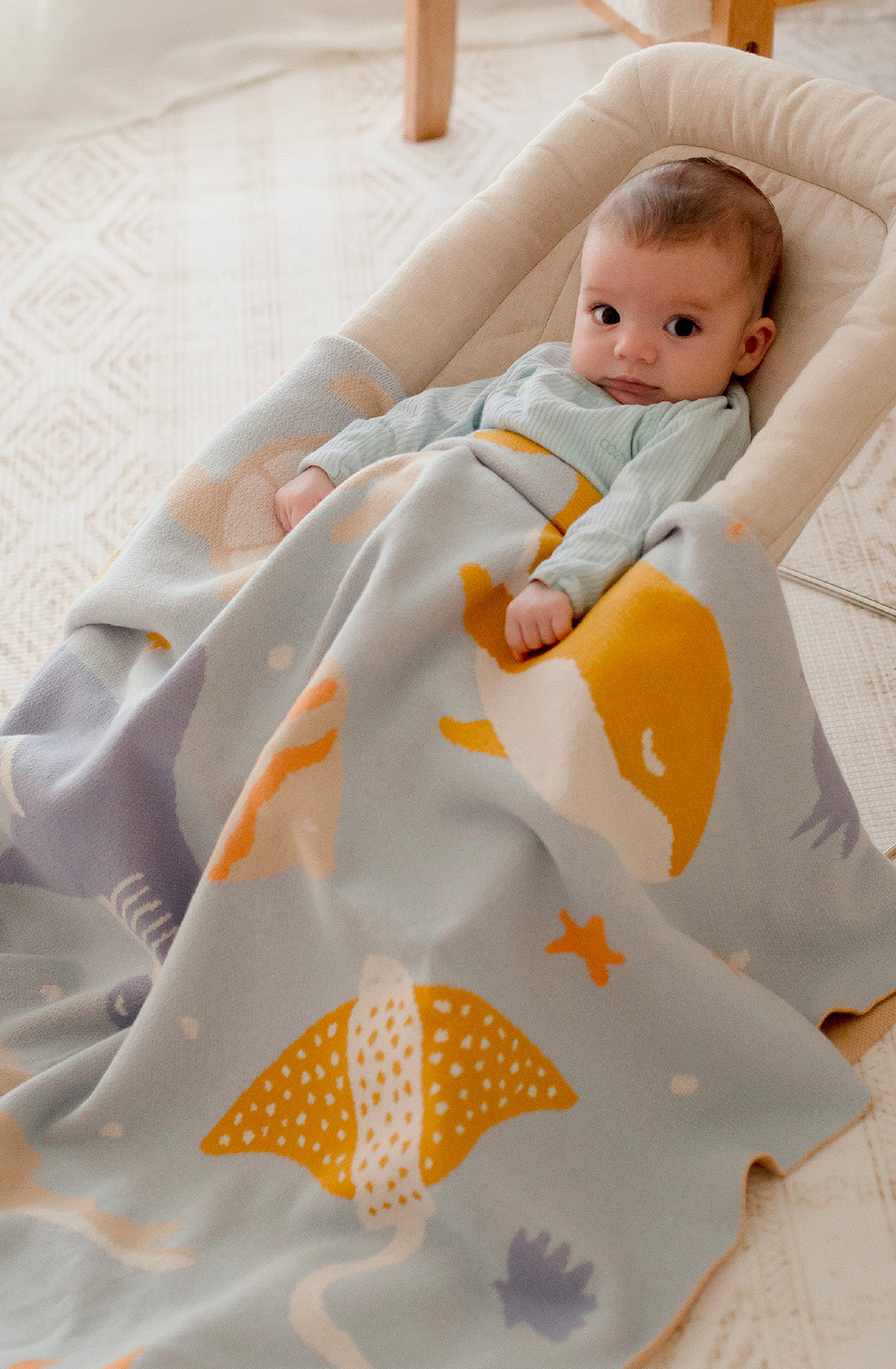 A baby lies in a cushioned infant lounger, covered with FOX & FALLOW's Ocean Creatures Blanket, adorned with colorful marine life illustrations, including a stingray and fish. The baby is dressed in a light green outfit and gazes toward the camera, surrounded by the cozy comfort of Oeko-Tex Standard 100 certified materials.