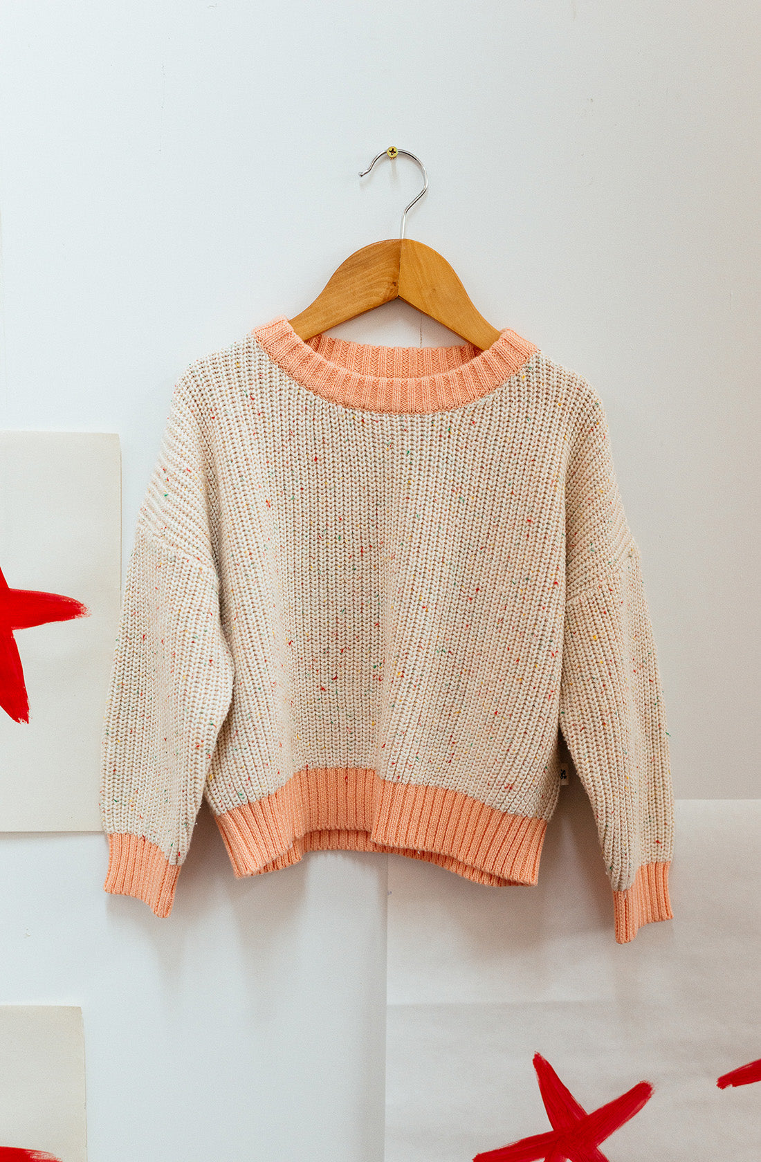 A Freckle Knit Peach jumper from SUNDAY SIBLINGS with pink trim is displayed on a wooden hanger against a white wall. Made from 100% cotton, the sweater features a chunky texture and speckled detailing for a comfortable fit. Red star-shaped illustrations are partially visible on either side of the sweater.